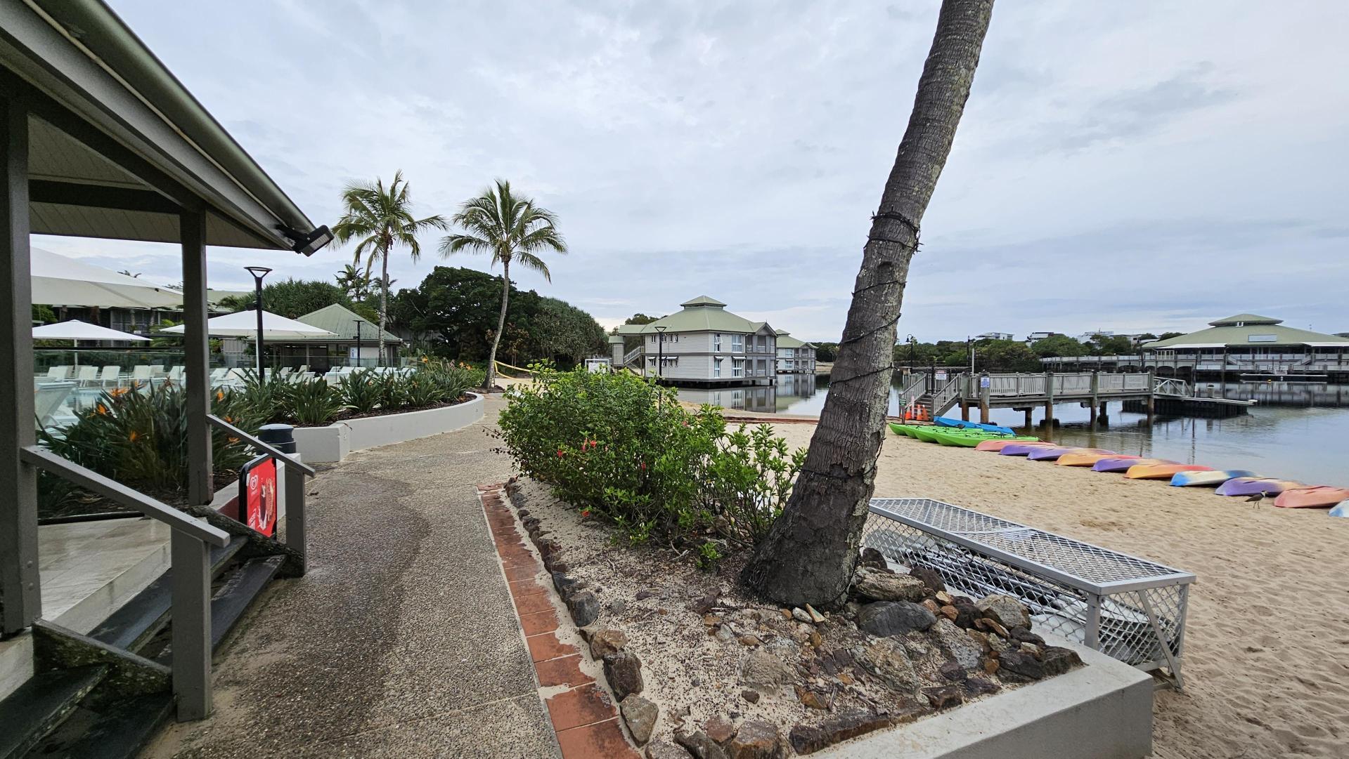 A sandy path curves along the left side, bordered by lush greenery and a building with a slanted roof. Two tall palm trees stand prominently in the centre. On the right, a sandy beach features a row of colourful kayaks near a calm body of water. A wooden pier extends over the water, leading to buildings on stilts. A metal bench is positioned near the beach, and the area is surrounded by tropical plants.