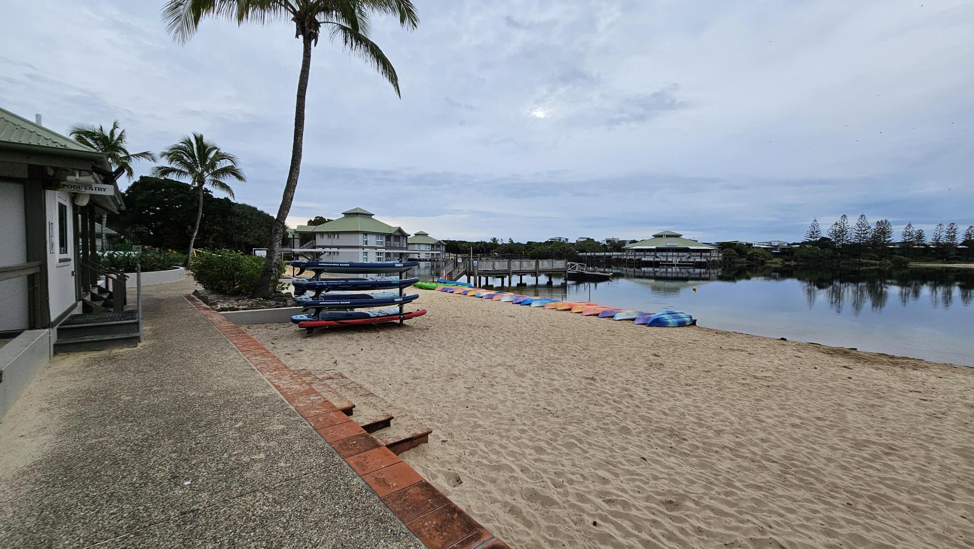 A sandy beach with a stack of surfboards on the left, next to a building with a green roof. A palm tree stands nearby. In the background, there are several buildings with green roofs and a wooden pier extending over calm water. A row of colourful kayaks is lined up along the shore. Trees are visible in the distance.