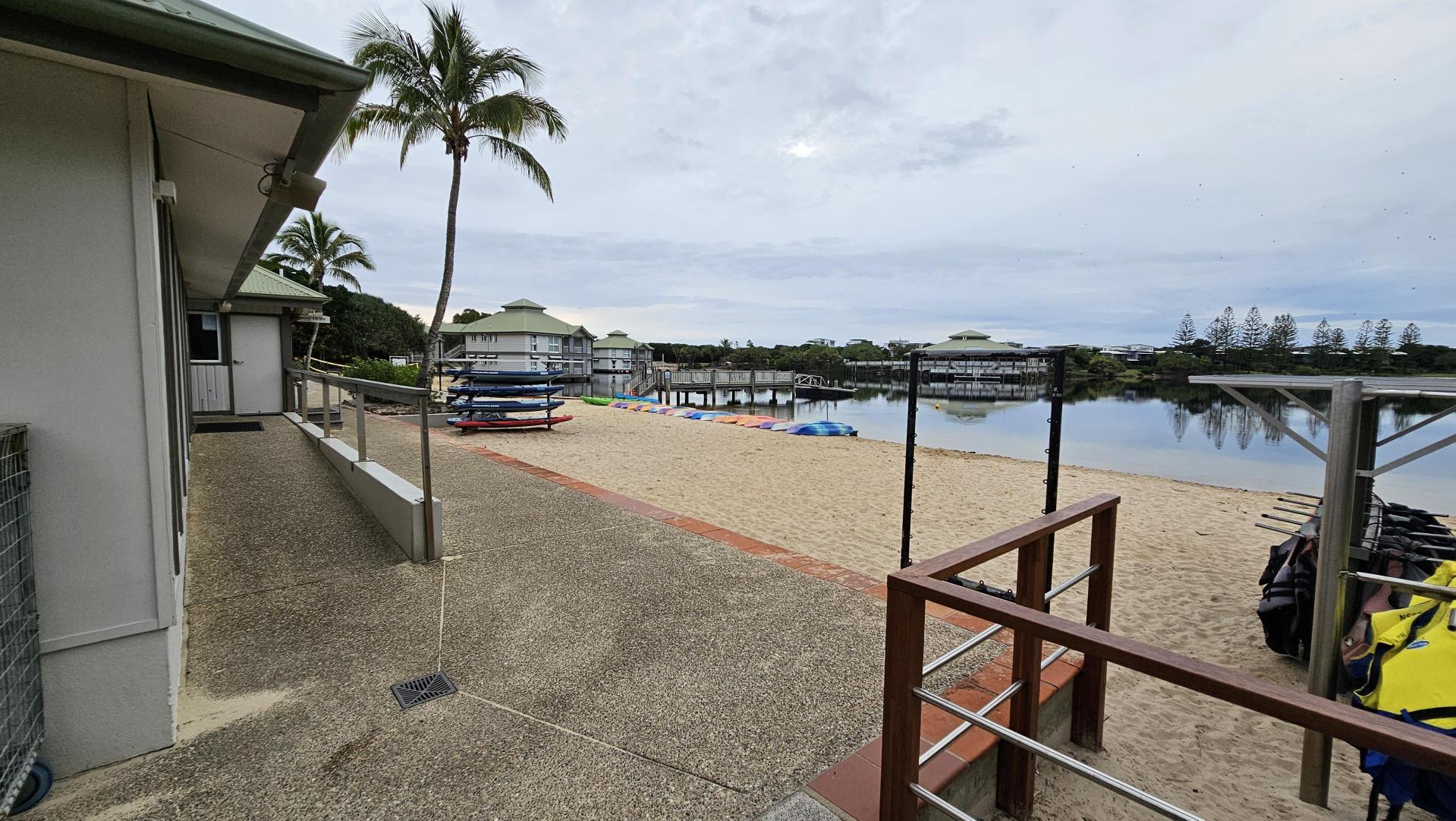 A sandy beach area with a stack of colourful kayaks and paddleboards on the left. A building with a green roof is adjacent to the beach, with a palm tree nearby. In the background, there are several waterfront houses on stilts, and a calm body of water reflecting the surroundings. On the right, there are racks with life jackets. The ground is a mix of sand and a paved walkway.