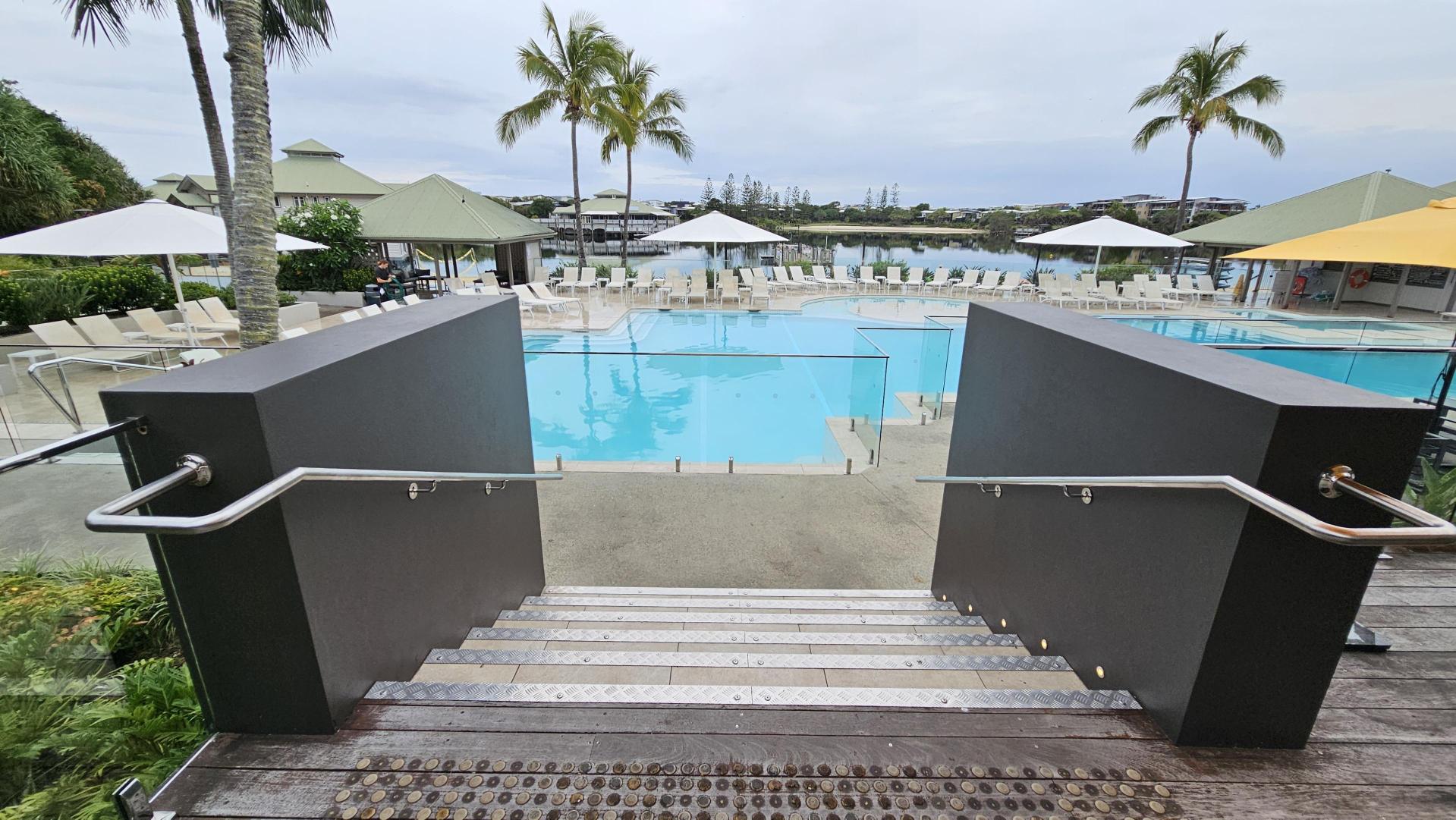 Steps lead down to a large swimming pool surrounded by sun loungers and umbrellas. Two palm trees are visible near the pool, and there are green-roofed structures in the background. The pool area is bordered by glass fencing, and the ground is a mix of wood and concrete. The scene is set in a landscaped area with greenery on the left.
