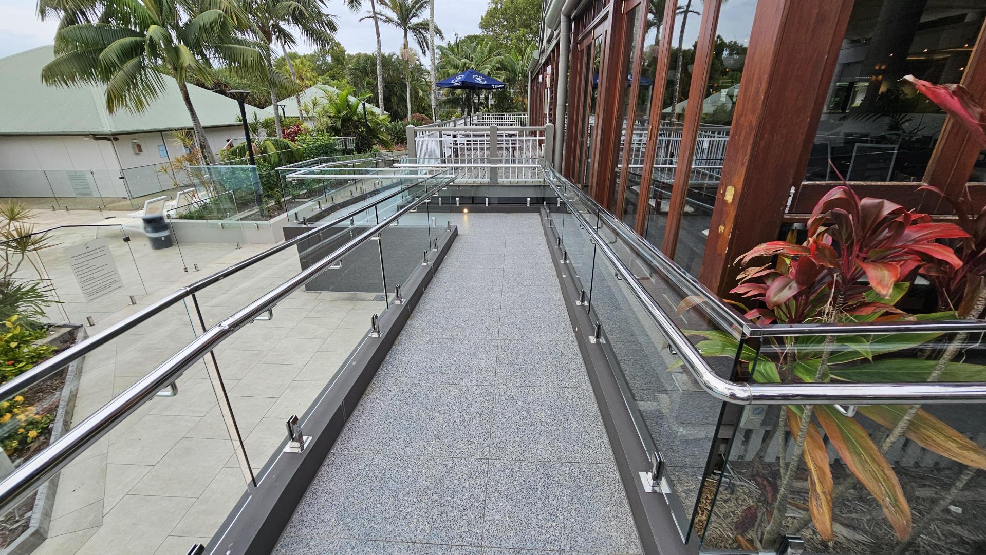 A walkway with a speckled grey floor and glass railings on both sides. To the left, there are vibrant plants and flowers next to a swimming pool with sun loungers. In the background, there are palm trees and a building with a green roof. On the right, a white metal fence runs parallel to the walkway.