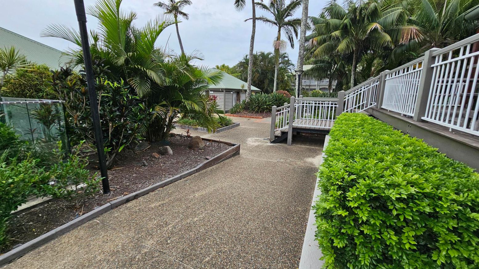 A pathway with a textured surface runs through a garden area. On the left, there are lush green plants and a black lamp post. On the right, a neatly trimmed hedge lines the path, with a white railing above it. In the background, tall palm trees and a small building with a green roof are visible. A wooden ramp with railings is on the right side of the path.