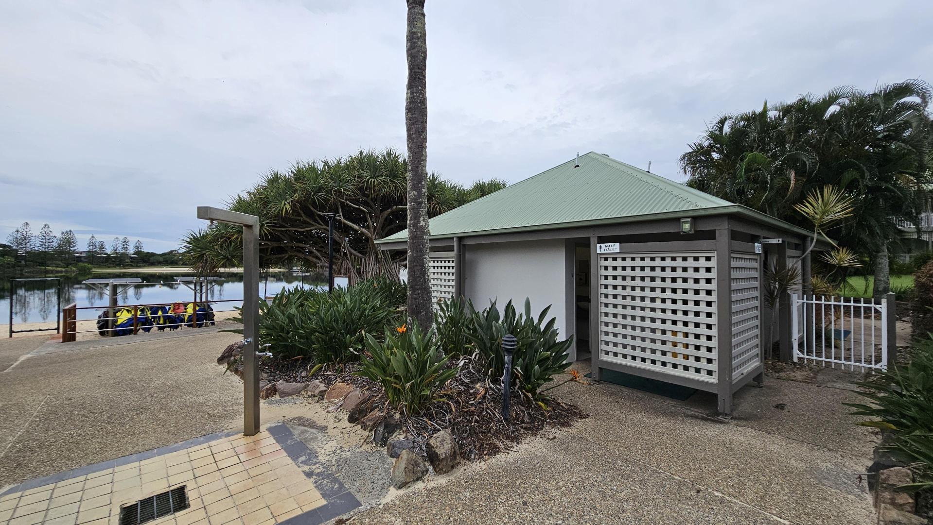 A small building with a green roof and lattice walls is on the right, surrounded by lush plants and a palm tree. In the foreground, there's a paved area with a metal shower fixture. On the left, a group of people in colourful clothing sit on a bench near a calm body of water. Trees and a fence line the background.
