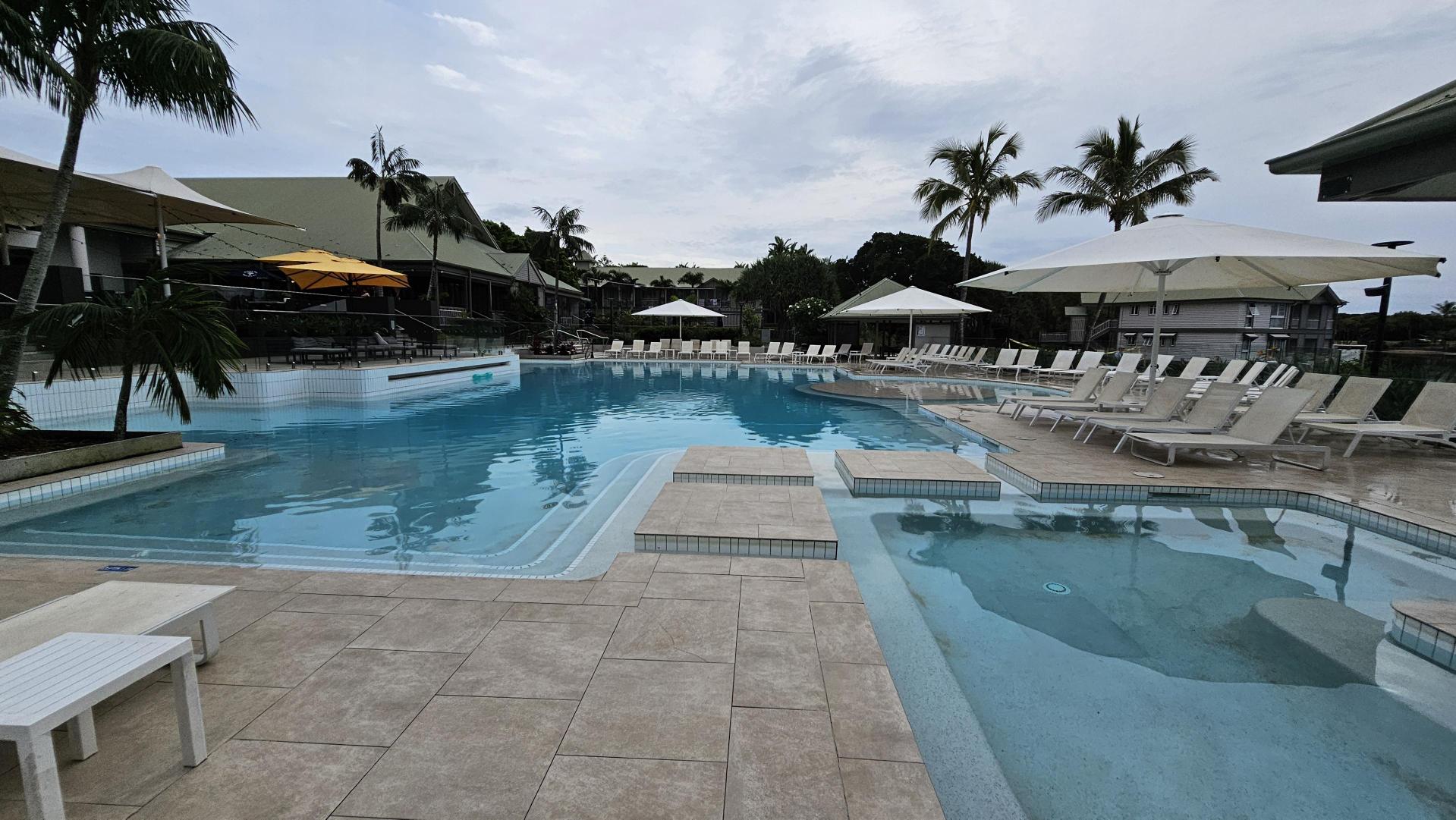A swimming pool area with light blue water, surrounded by beige tiled flooring. On the right, there are numerous white sun loungers under large white umbrellas. In the background, palm trees and a building with a sloped roof are visible. On the left, a yellow umbrella is seen near more seating. The pool has a stepped entrance and a small raised platform in the water.