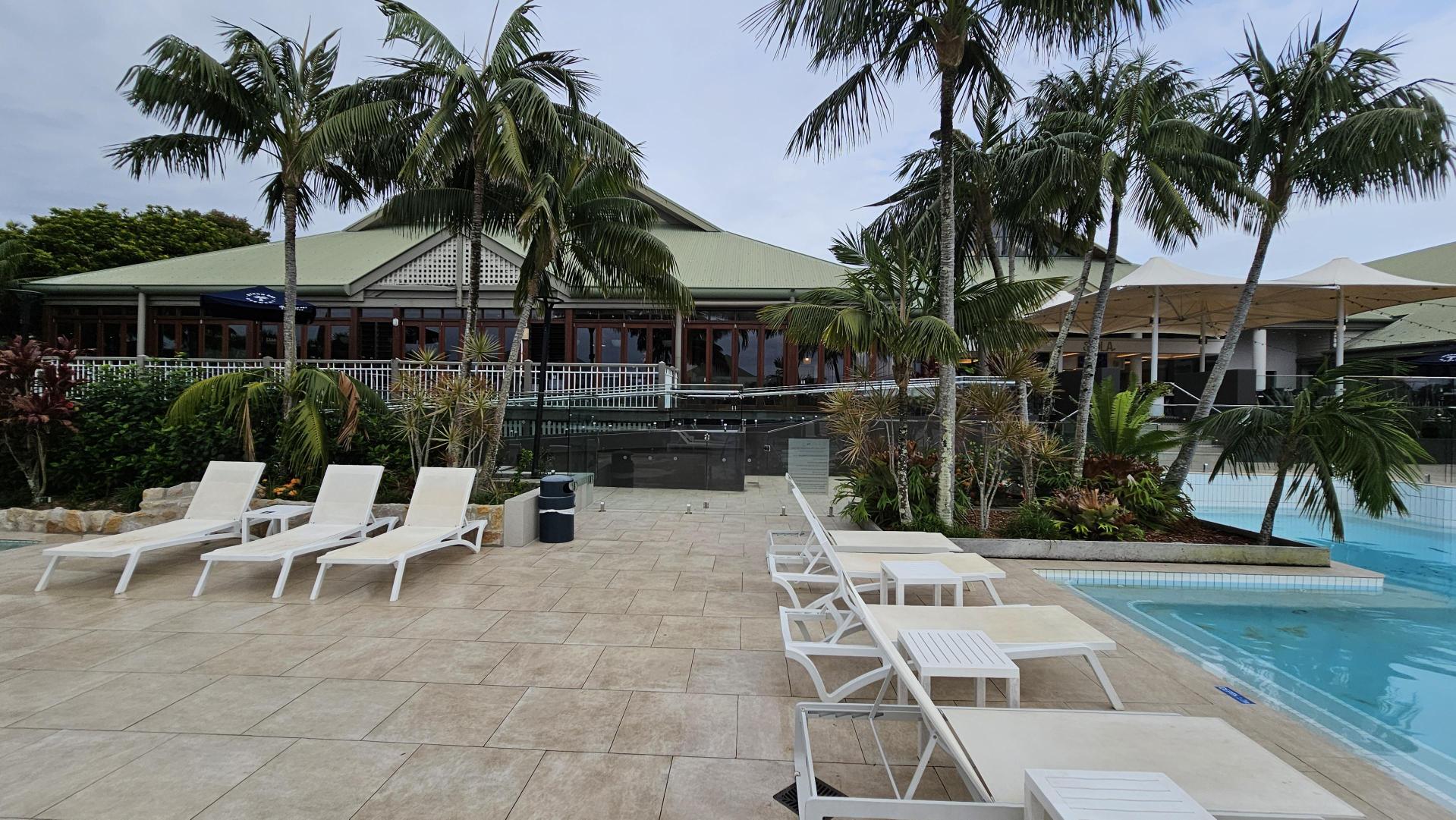 Poolside area with several white sun loungers and small tables on a tiled patio. To the right, a clear blue swimming pool is visible. In the background, there are tall palm trees and a large building with a green roof and red-framed windows. The building is surrounded by lush greenery and has a white railing.