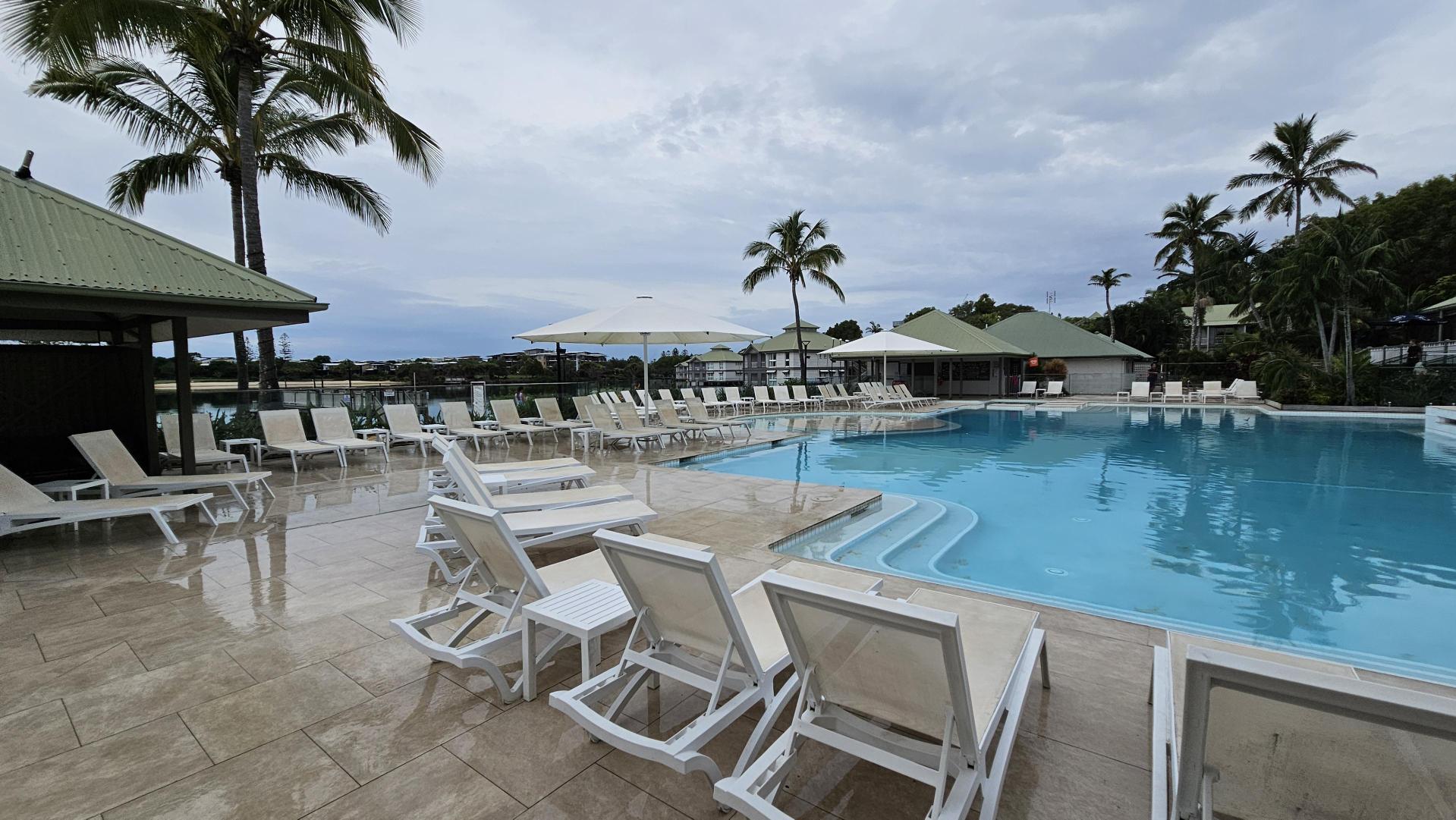A swimming pool area with light blue water is surrounded by white sun loungers and tables. On the left, there is a structure with a green roof and a tall palm tree. In the background, more palm trees and buildings with green roofs are visible. The pool deck is made of light-coloured tiles, and there are large white umbrellas providing shade.