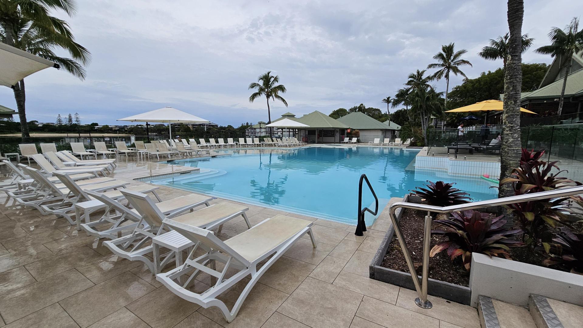 A swimming pool surrounded by white sun loungers and large umbrellas. On the left, several loungers are lined up on a tiled floor. In the centre, the pool is clear and inviting, with a metal handrail leading into the water. On the right, there are more loungers and a small garden area with tropical plants. In the background, palm trees and a building with a green roof are visible.