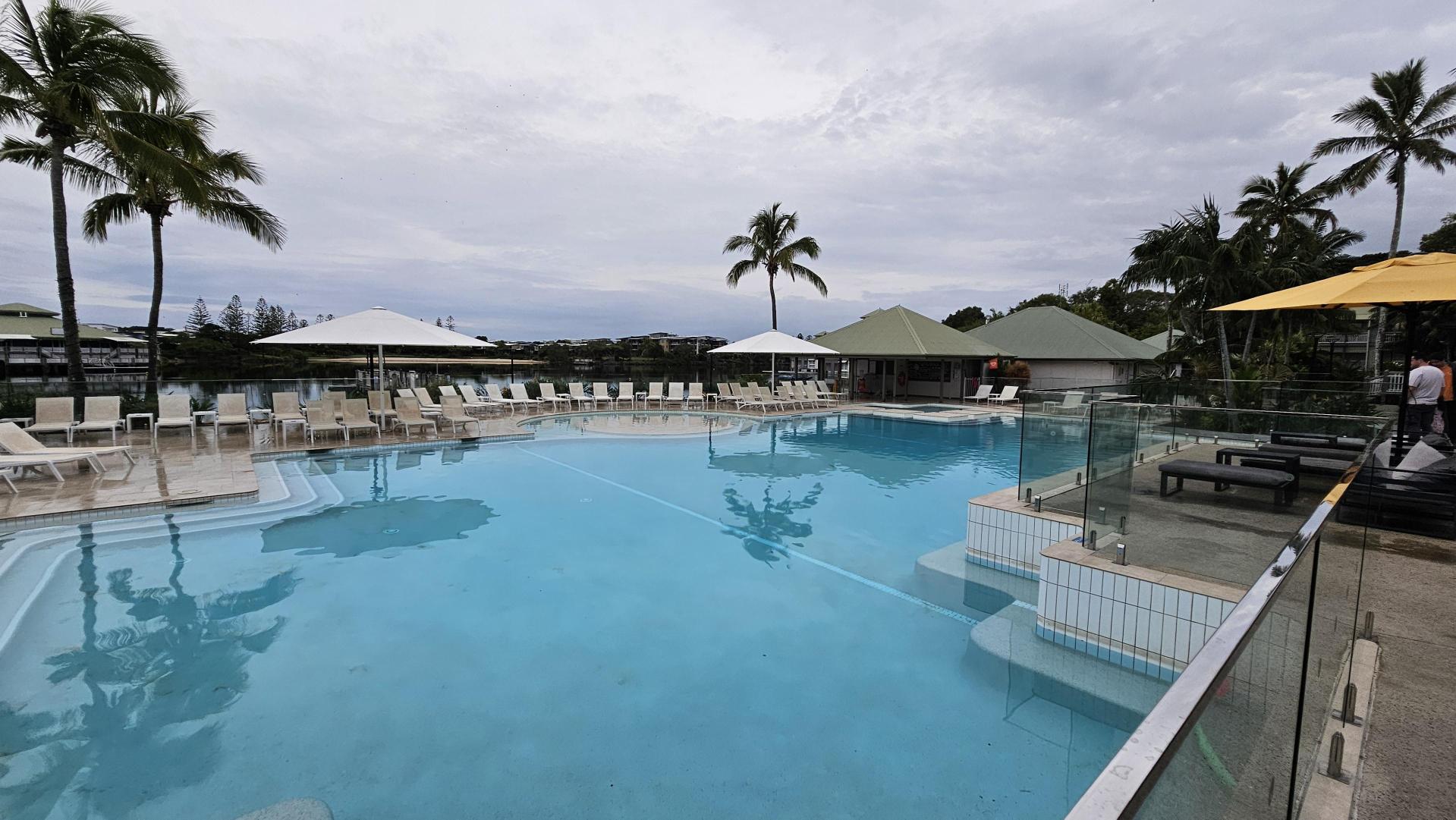 A large outdoor swimming pool with clear blue water is surrounded by white sun loungers and large umbrellas. On the left, there are tall palm trees. On the right, a glass barrier separates the pool from a seating area with dark cushioned benches under a yellow umbrella. In the background, there are more palm trees and a building with a green roof. The pool area is spacious and well-maintained.