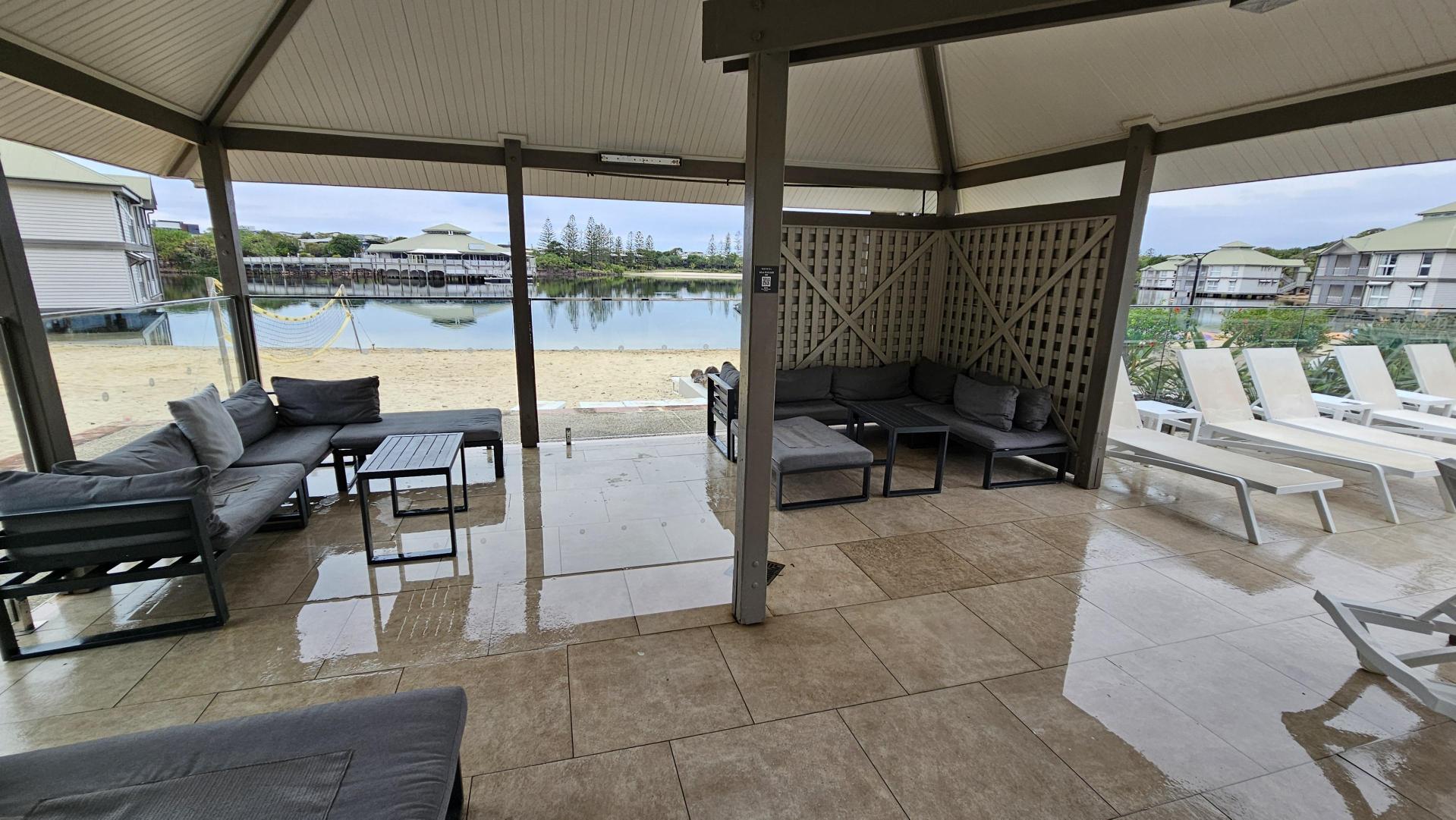 A covered patio with a slanted beige ceiling and tiled floor. On the left, grey cushioned seating with small black tables faces the centre. On the right, more grey cushioned seats are arranged in an L-shape against a lattice-panelled wall, with white pool loungers beyond. The background features a sandy area, a glass barrier, and a still body of water reflecting surrounding structures and trees.