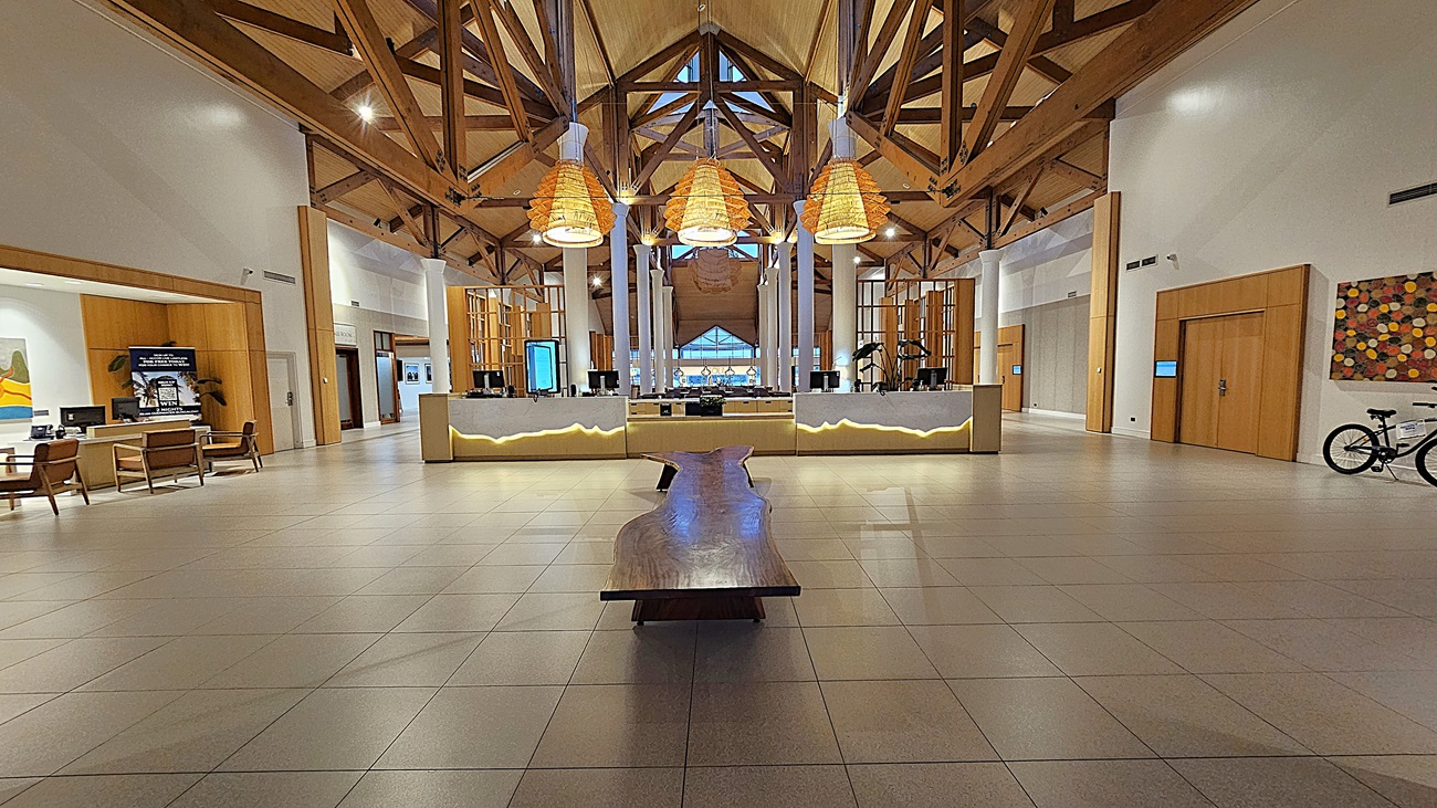 On the left, there are seating areas with wooden chairs and tables, and a digital display screen mounted on the wall. To the right, a bicycle is parked against the wall, and a colourful abstract painting is hung nearby. In the middle, a long wooden bench extends towards the reception desk, which is illuminated with soft lighting. The floor is tiled in light grey, and the ceiling features exposed wooden beams and large, woven light fixtures.