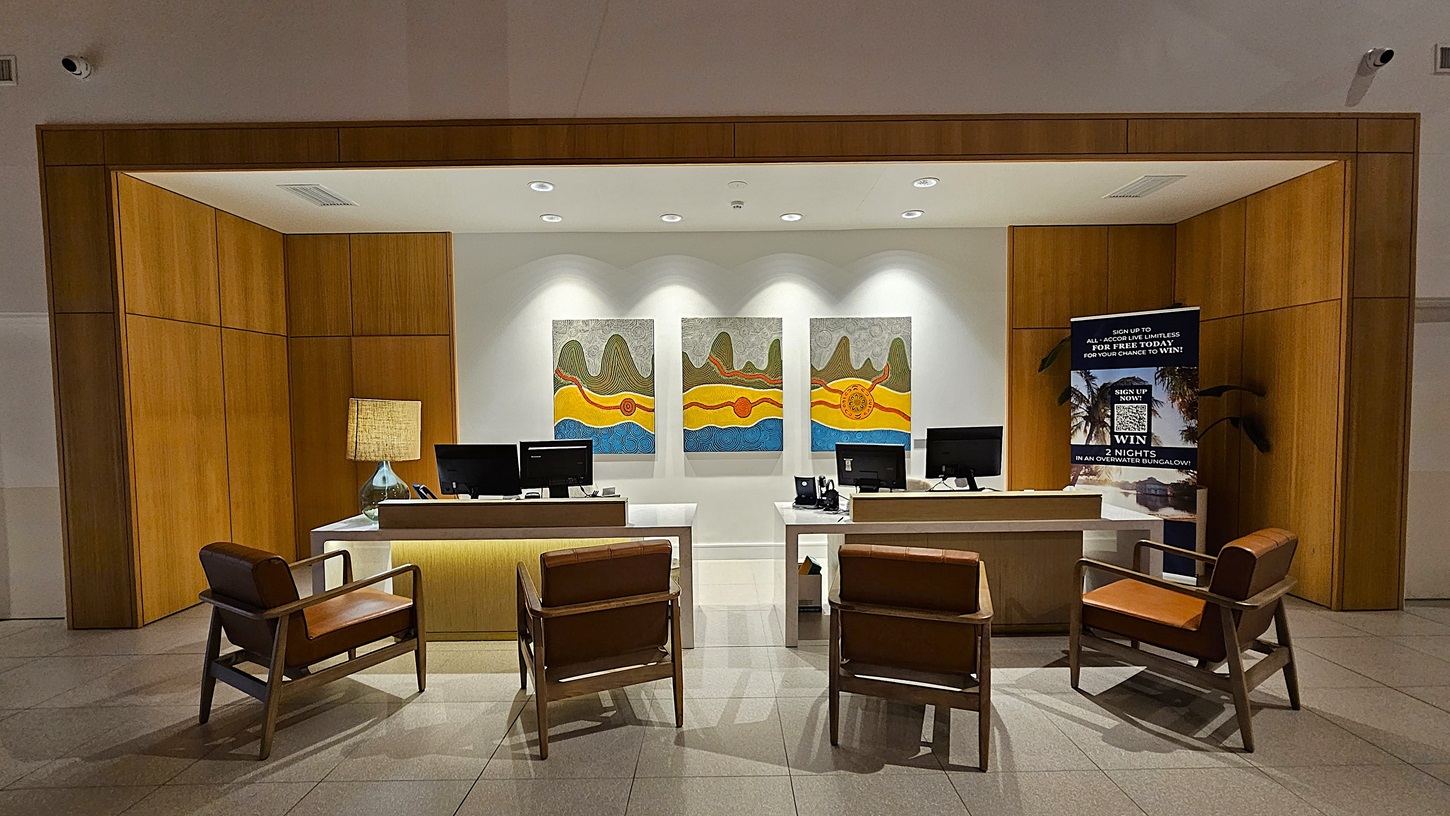On the left, there is a wooden panelled wall with a table lamp on a desk. To the right, a large promotional sign stands next to another desk. In the middle, three colourful abstract paintings hang on a white wall above two desks equipped with computer monitors and office supplies. Four brown leather chairs are arranged in front of the desks. The floor is tiled in light grey, and the ceiling is white with recessed lighting, creating a well-lit workspace.
