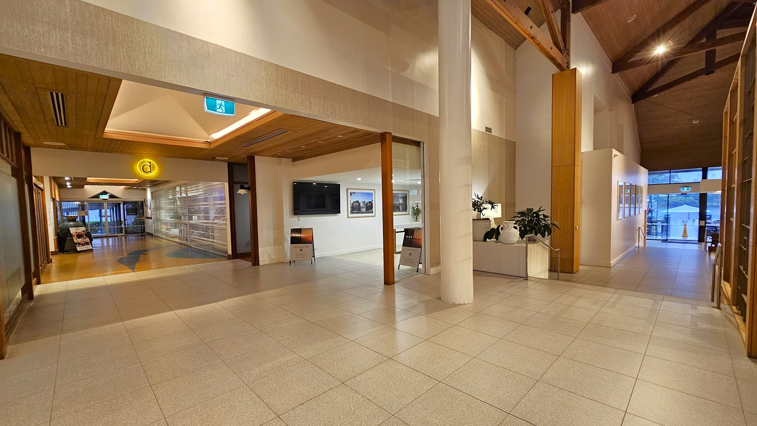 On the left, there is a hallway with a wooden ceiling and a yellow circular sign. To the right, a white column stands next to a planter with green plants and a white vase. In the middle, a large open space with light grey tiled flooring leads to a reception area with a black screen on the wall and two signs below it. The ceiling is high with exposed wooden beams and recessed lighting, creating a spacious and well-lit environment.