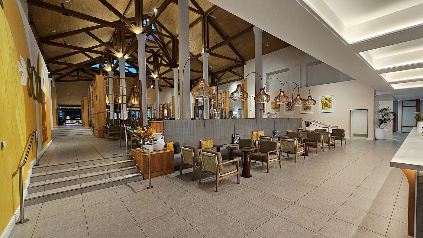 On the left, a yellow wall with a decorative mural and a metal handrail leads up a few steps to a higher level. To the right, a seating area with wooden chairs and small tables is arranged under large, woven pendant lights. In the middle, a grey partition wall separates the seating area from the rest of the space. The floor is tiled in light grey, and the ceiling is high with exposed wooden beams and recessed lighting, creating an open and well-lit environment.
