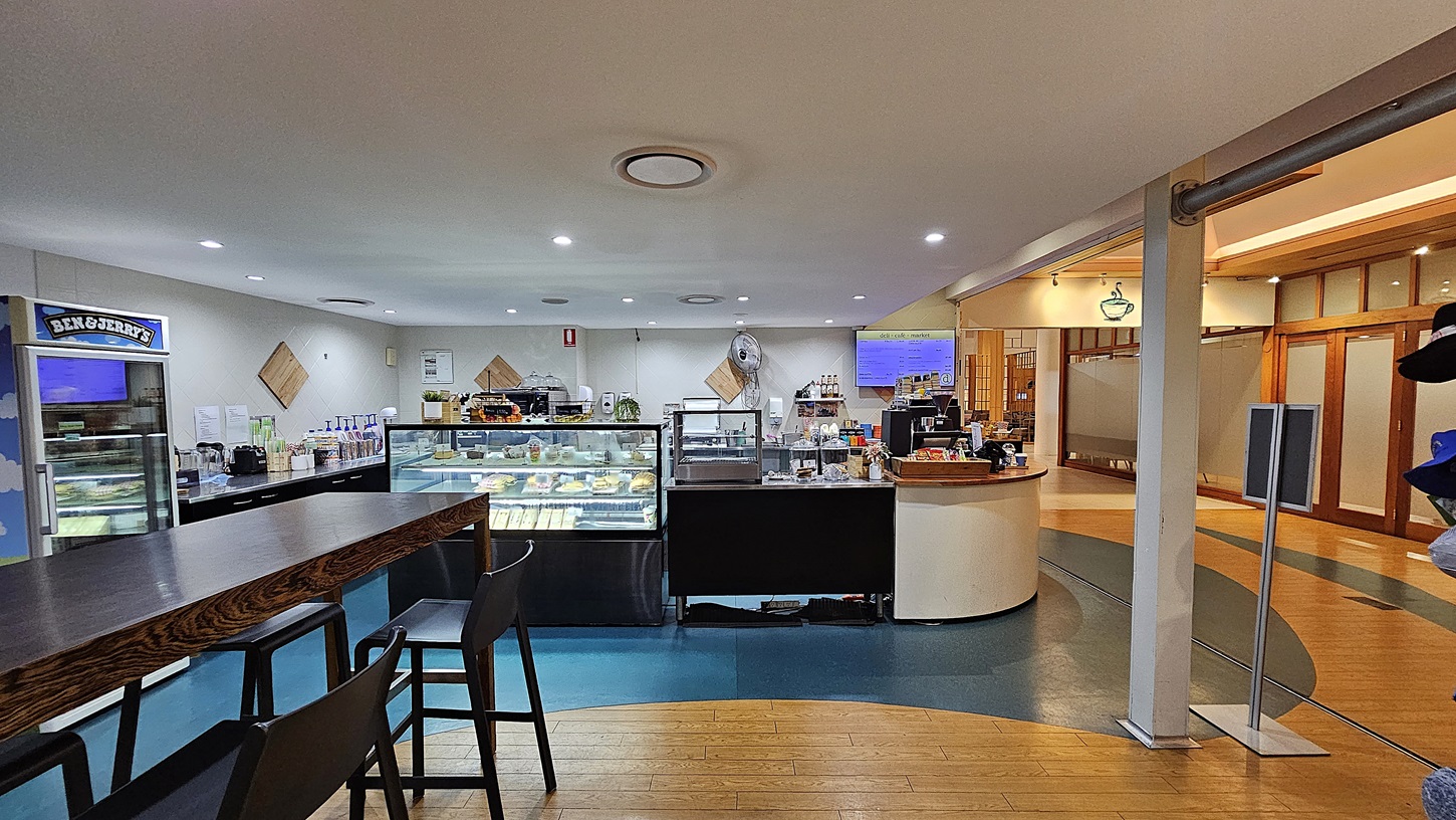 On the left, there is a Ben & Jerry's freezer next to a long wooden table with high chairs. To the right, a curved counter with a cash register and various items leads to a hallway with wooden doors and frosted glass panels. In the middle, a display case filled with pastries and a coffee machine is set against the back wall. The floor transitions from wooden planks to blue linoleum, and the ceiling is white with recessed lighting and circular vents.