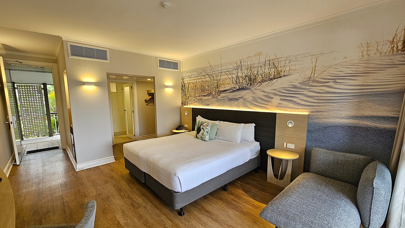 On the left, an open door leads to a balcony with a lattice screen and greenery. To the right, a grey upholstered chair and a round wooden side table. In the middle, a large bed with white linens, decorative pillows, and a beach-themed mural on the wall behind it. The floor is wooden, and the ceiling is white with recessed lighting and air vents.