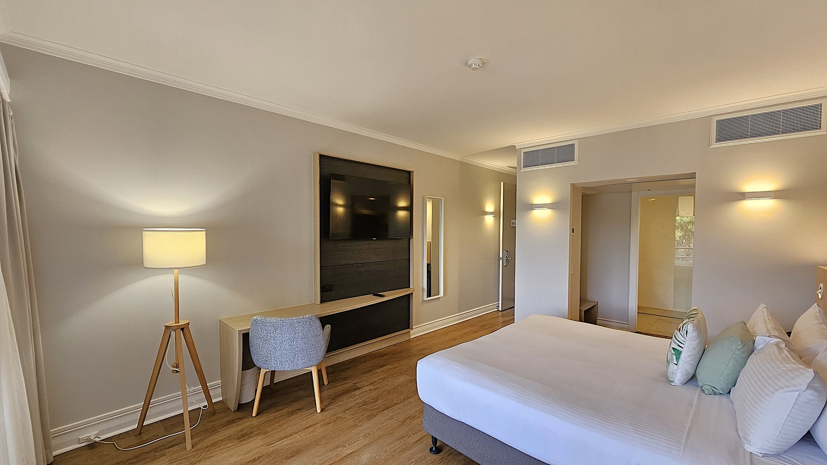On the left, a wooden tripod floor lamp with a beige shade and a grey upholstered chair at a wooden desk. To the right, a large bed with white linens and decorative pillows. In the middle, a wall-mounted flat-screen TV above the desk and a full-length mirror next to the entrance door. The background features an open doorway leading to a tiled area. The floor is wooden, and the ceiling is white with recessed lighting and air vents.