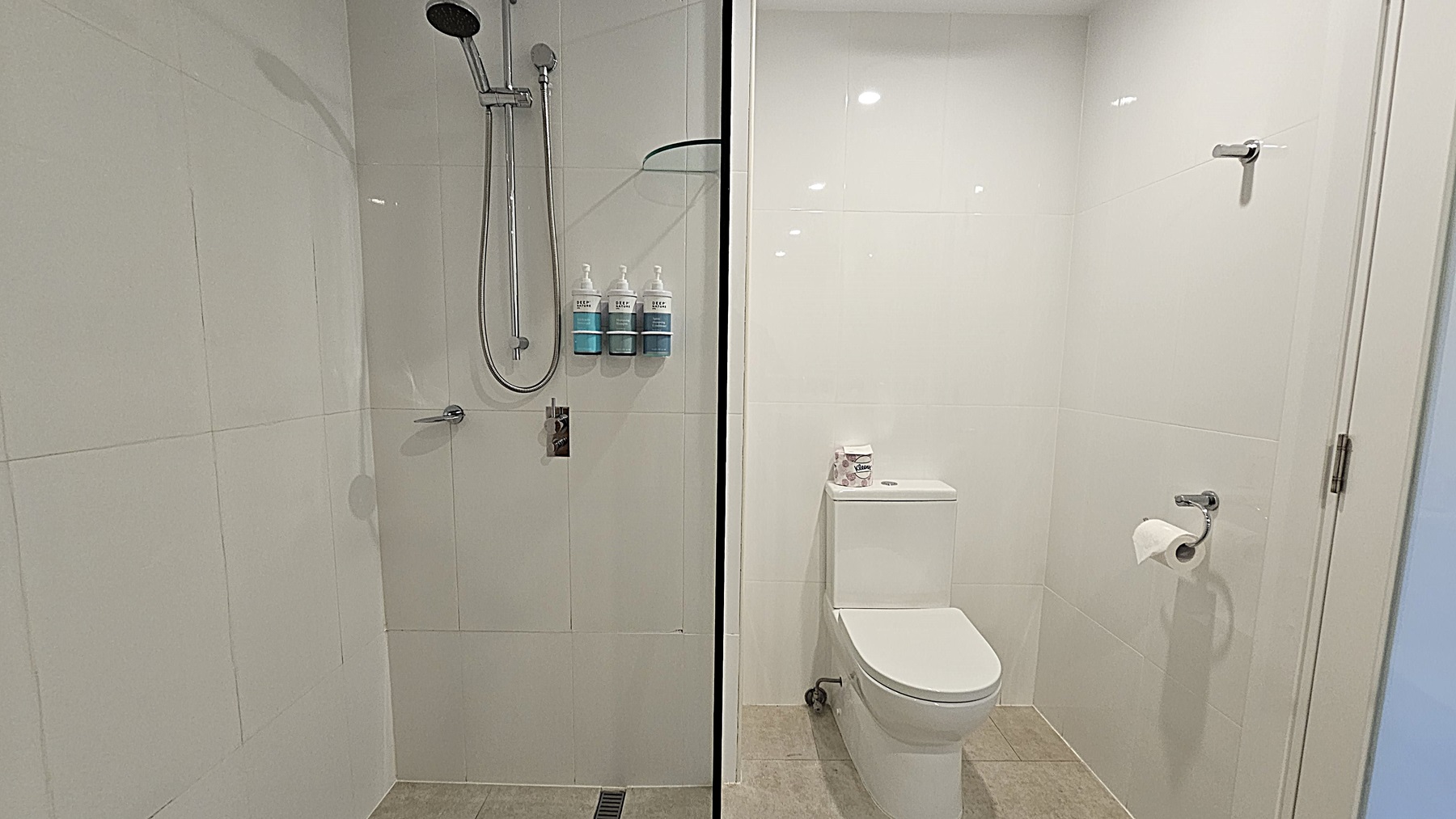 On the left, a shower area with a wall-mounted showerhead, a handheld shower, and three soap dispensers. To the right, a white toilet with a roll of toilet paper on a holder and a small shelf above it holding additional toilet paper rolls. The floor is tiled in light grey, and the ceiling is white with recessed lighting.