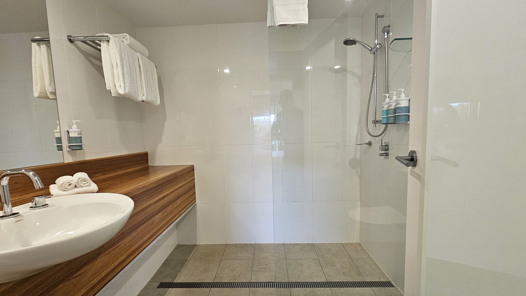 On the left, a white sink with a chrome faucet set into a wooden countertop, above which are two soap dispensers and a towel rack holding folded white towels. To the right, a glass-enclosed shower area with a wall-mounted showerhead, a handheld shower, and three soap dispensers. The floor is tiled in light grey, and the ceiling is white with recessed lighting.