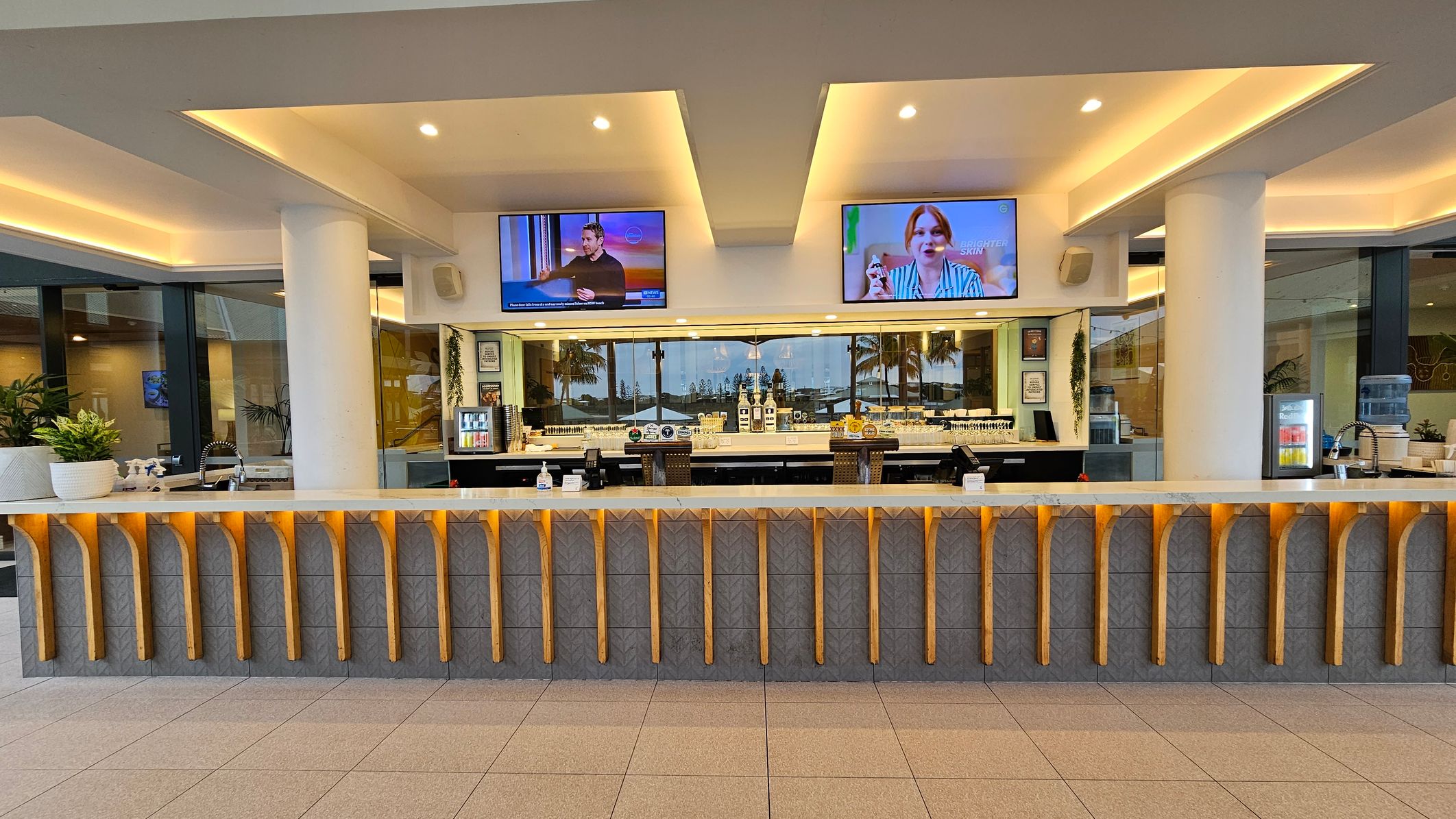 Spacious interior with a high, wooden-beamed ceiling. On the left, a yellow wall with artwork and a staircase with a metal railing. In the centre, a seating area with wooden chairs, tables, and a grey partition. Above, large, decorative pendant lights. On the right, a long counter and a white wall with framed art. The floor is tiled, and there are potted plants near the walls.
