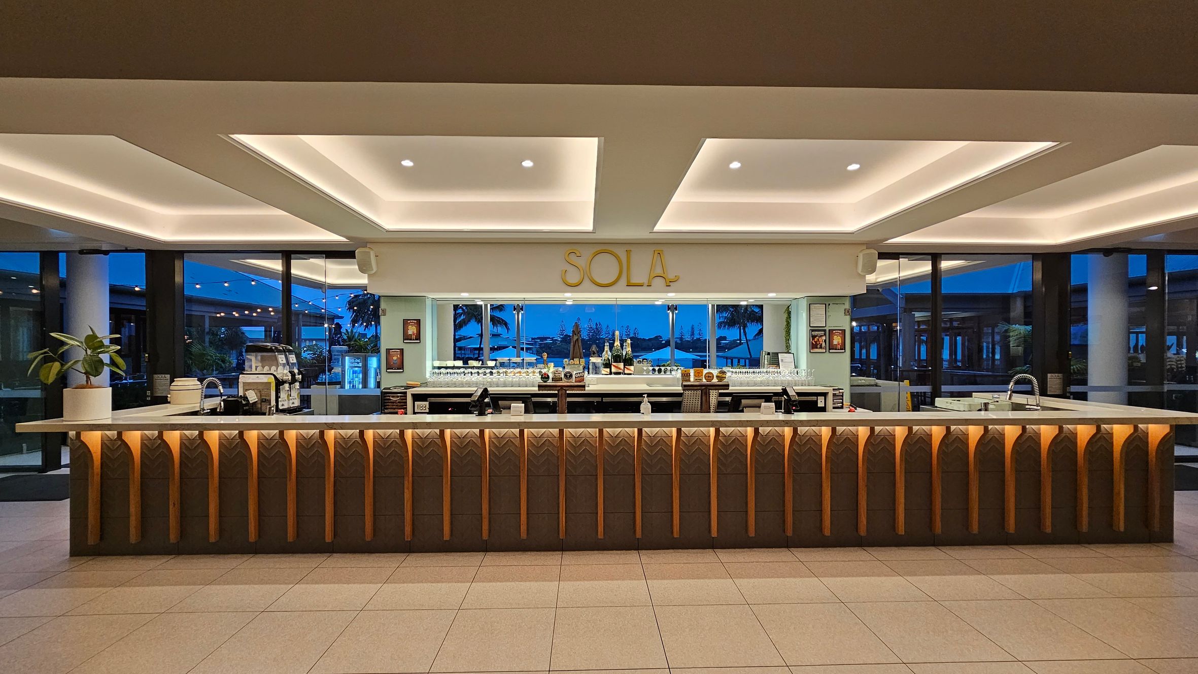 A modern bar area with a long counter featuring geometric patterns and warm lighting. Behind the counter, there are shelves with bottles and glasses, and a sign reading "SOLA" above. Large windows in the background reveal an outdoor view with palm trees and a blue sky. The ceiling has recessed lighting, and the floor is tiled. A plant is placed on the left side of the counter.