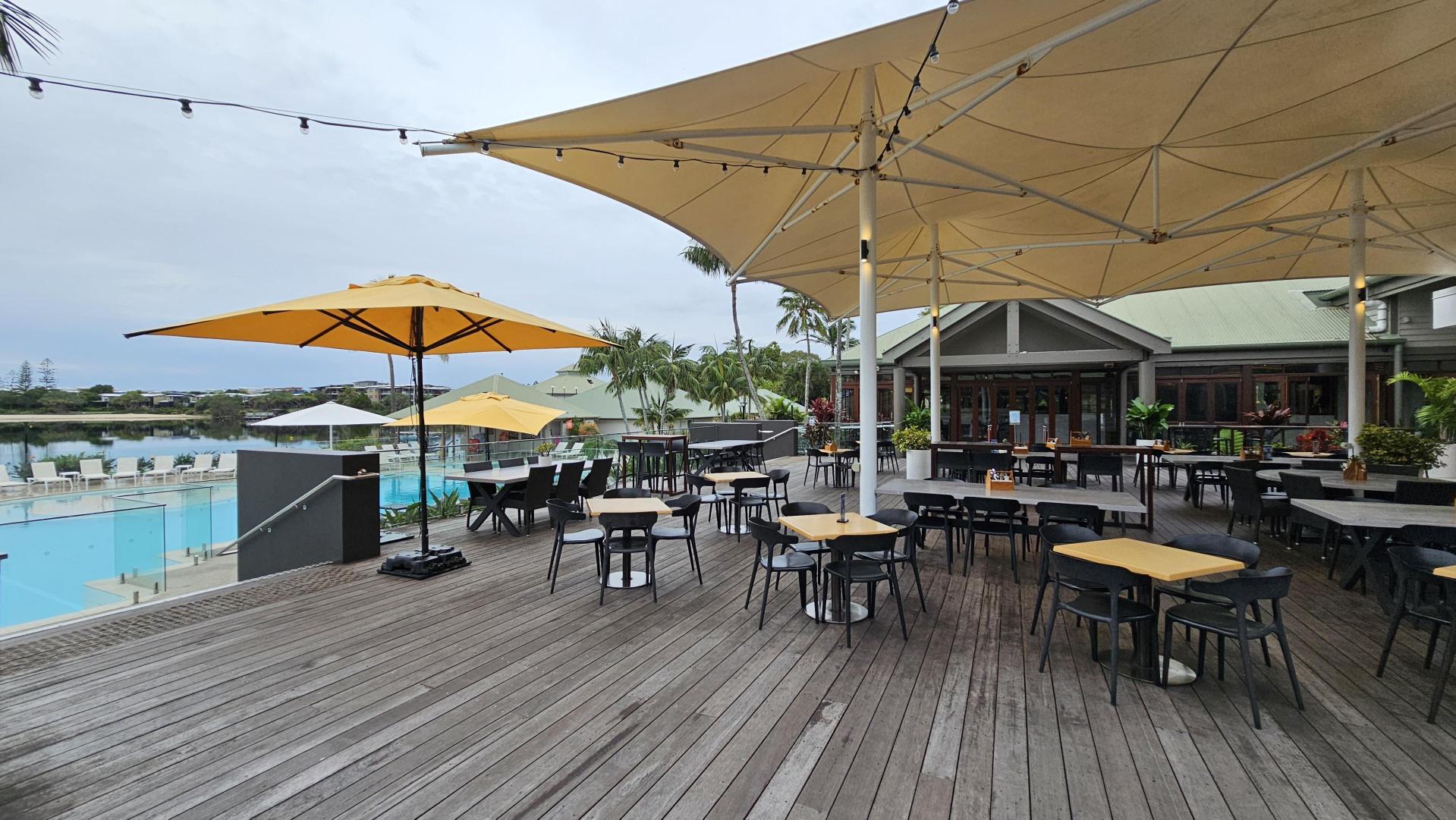 Outdoor dining area with wooden decking and several tables and chairs. Large yellow umbrellas provide shade. In the background, there's a swimming pool with sun loungers and a glass railing. A building with a green roof is visible, surrounded by palm trees and plants. Overhead, string lights are hung across the area.