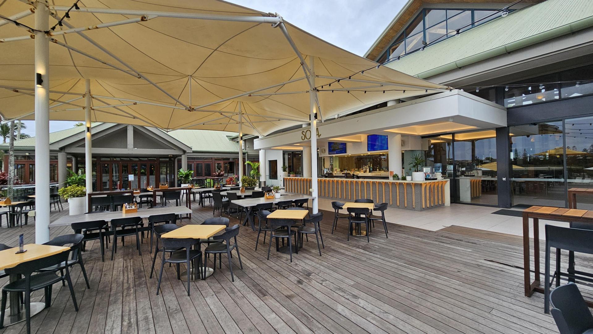 Outdoor dining area with wooden decking and several tables and chairs. Large beige umbrellas provide shade. On the right, there's a modern bar with a wooden counter and glass doors leading inside. In the background, a building with a peaked roof and large windows. Potted plants are scattered around, adding greenery to the space.