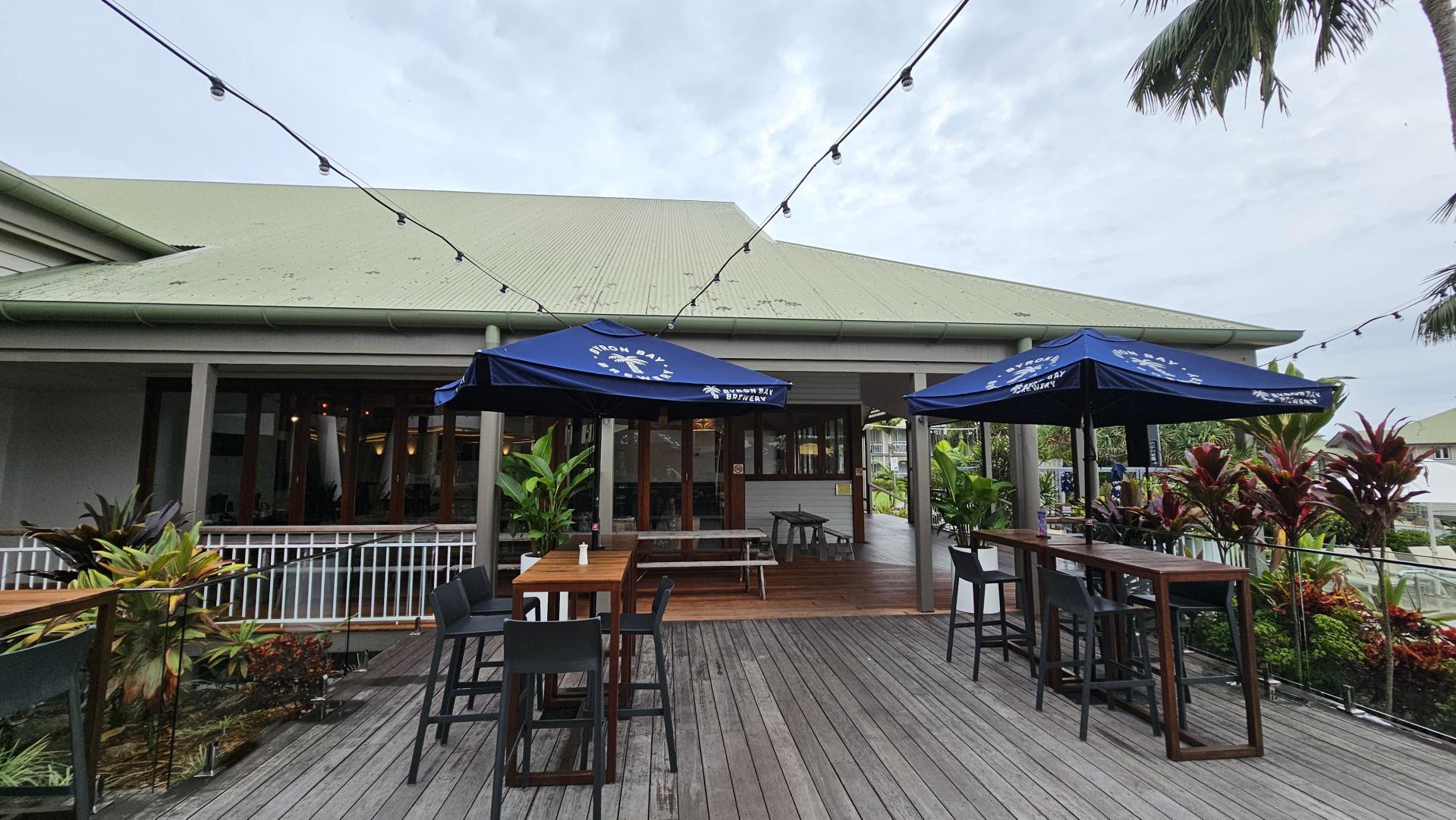 Outdoor seating area with wooden tables and black chairs on a wooden deck. Two blue umbrellas provide shade. String lights are hung above. The background features a building with a light green roof and large windows. Lush green plants and red-leaved shrubs line the perimeter. A palm tree is visible on the right.