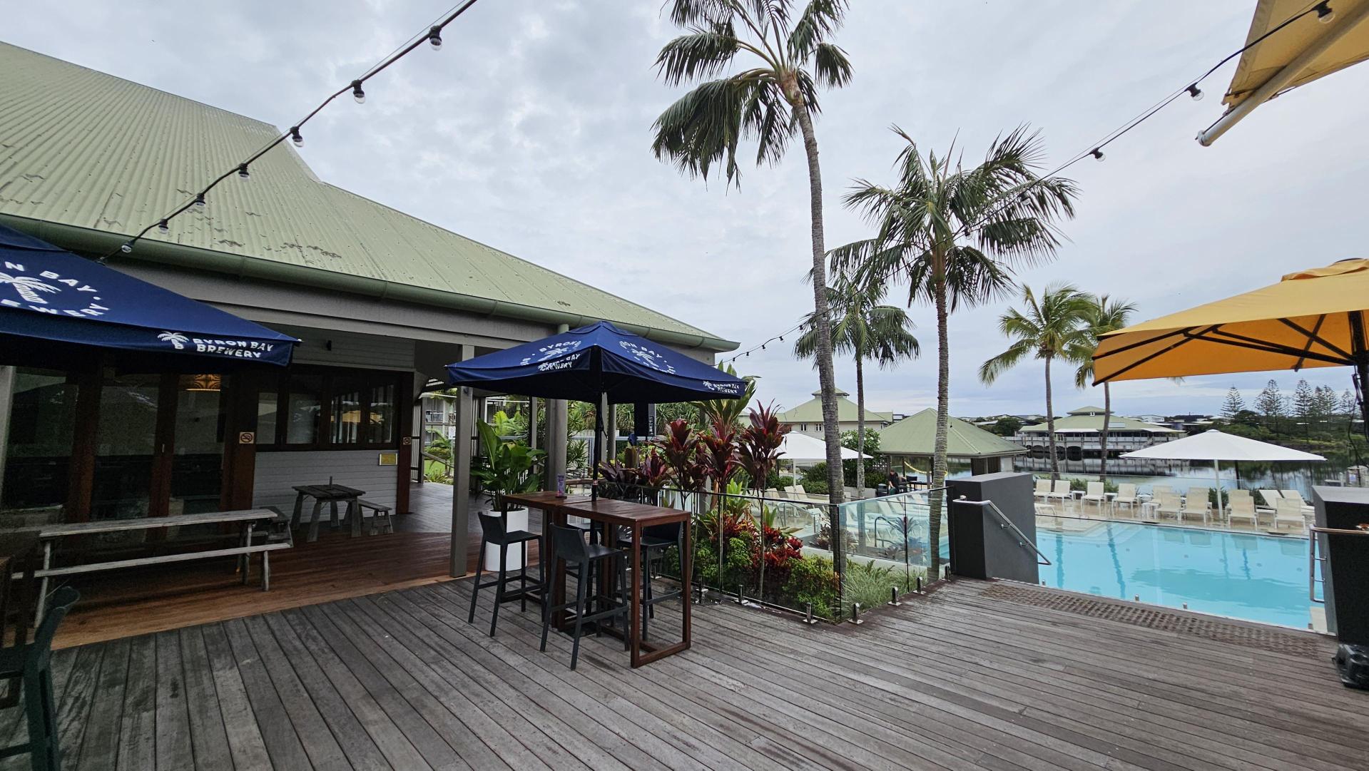 A wooden deck with outdoor seating, including tables and chairs under blue and yellow umbrellas. On the left, there's a building with a green roof and a sign on the umbrella. In the middle, potted plants and palm trees add greenery. On the right, a swimming pool is surrounded by white loungers and umbrellas. String lights are hung above, and the area has a relaxed, open-air atmosphere.