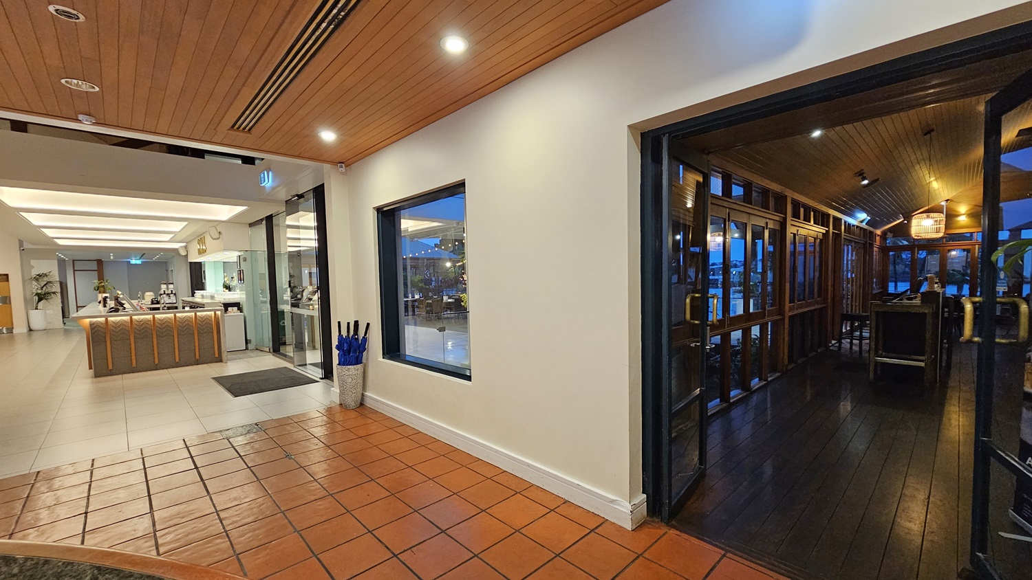 On the left, a staircase with a metal handrail leads to an elevated seating area with wooden partitions and potted plants. To the right, a long counter with a light-coloured surface extends towards the back, with a potted plant near the end. In the middle, a seating area with grey upholstered chairs and wooden tables is illuminated by hanging pendant lights. The floor is tiled in light grey, and the ceiling features exposed wooden beams and recessed lighting.