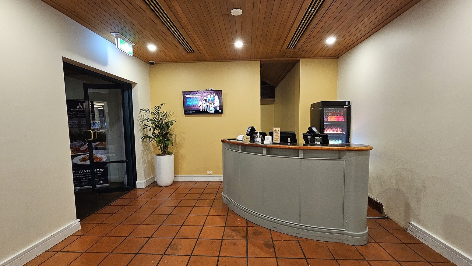 On the left, a black-framed glass door leads to another room, with a large potted plant next to it. To the right, a curved reception desk with a wooden top holds several electronic devices and a small display stand. In the background, a black refrigerator with illuminated shelves is against a yellow wall. The floor is covered in terracotta tiles, and the ceiling is wooden with recessed lighting and ventilation panels.