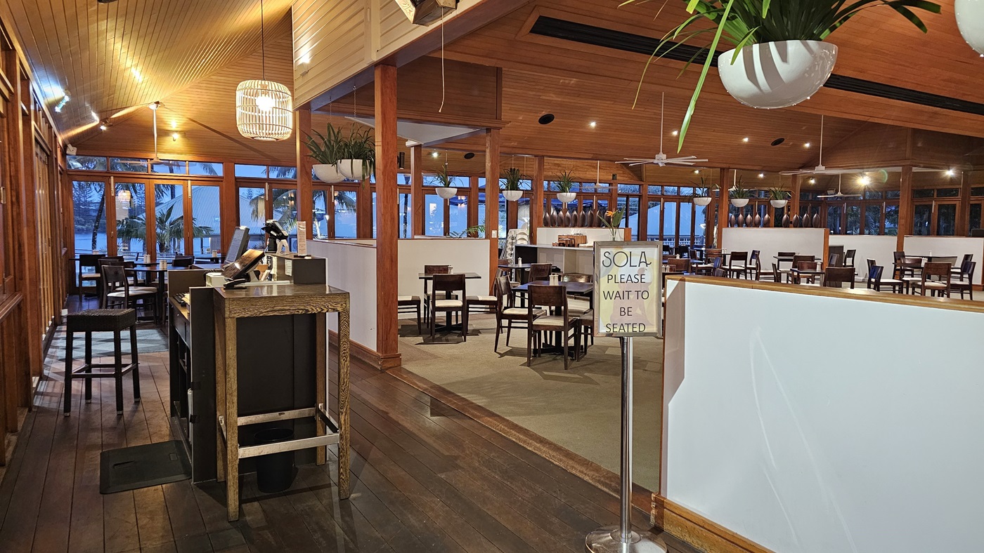 On the left, a wooden reception desk with a computer and a high stool is positioned near a wall of large windows. To the right, a sign reading "SOLA PLEASE WAIT TO BE SEATED" stands next to a white partition. In the background, multiple dining tables and chairs are arranged on a carpeted area, with hanging plants and pendant lights above. The floor is a mix of dark wood and carpet, and the ceiling is wooden with recessed lighting and ceiling fans.