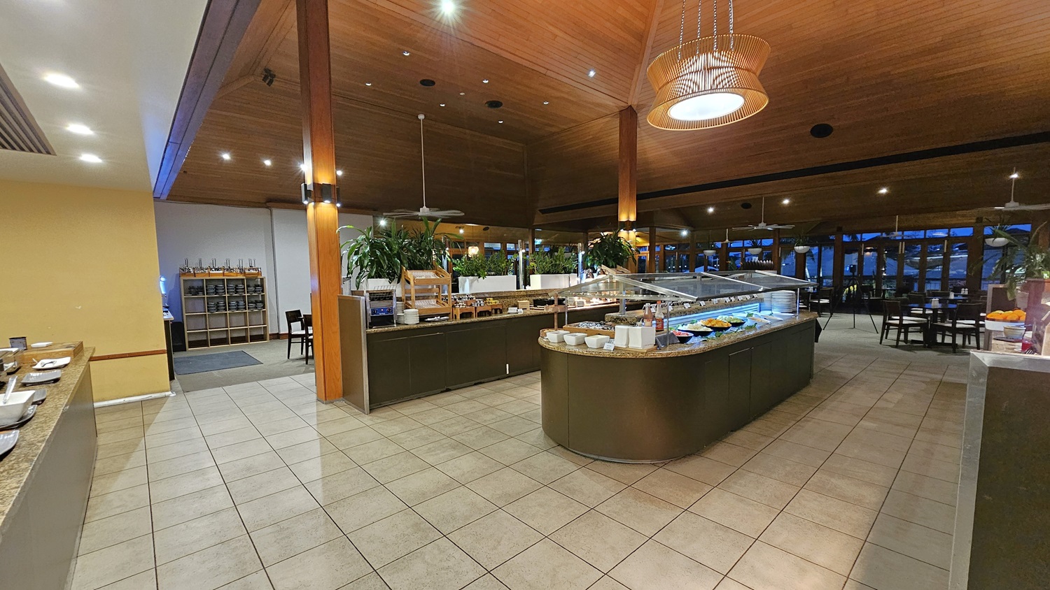 On the left, a granite counter with plates and bowls is positioned against a yellow wall. To the right, a large buffet counter with various food items and a glass sneeze guard extends towards the back. In the middle, another buffet counter with salads and condiments is surrounded by potted plants. The background features dining tables and chairs near large windows. The floor is tiled in light grey, and the ceiling is wooden with recessed lighting and a large pendant light.