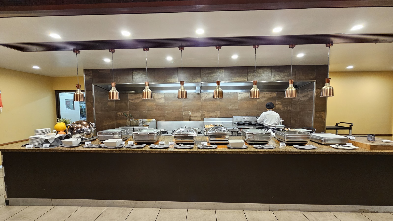 On the left, a stack of plates and bowls is arranged next to a fruit display on a granite counter. To the right, more serving dishes and utensils are placed on the same counter. In the middle, several chafing dishes with lids are lined up, with a chef working at a stove in the background. The floor is tiled in light grey, and the ceiling is white with recessed lighting and copper pendant lights hanging above the counter.