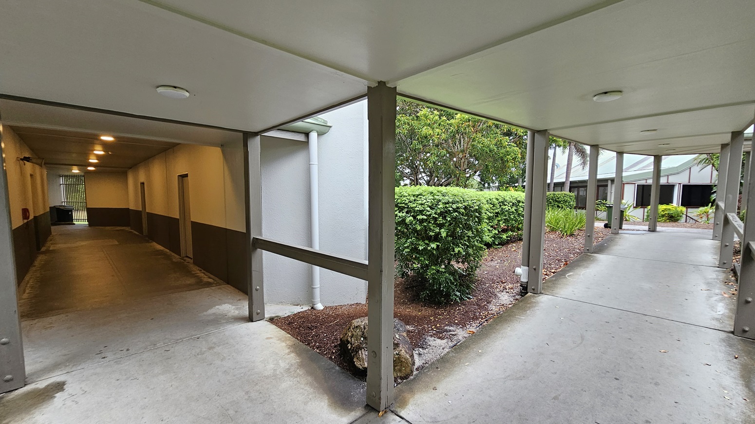 On the left, an enclosed corridor with beige walls and a grey lower section has doors and ceiling lights. To the right, an open walkway with grey pillars and a concrete floor leads past green bushes and trees. In the middle, a garden area with shrubs and a tree is visible. The floor is concrete, and the ceiling is flat and white with recessed lighting.