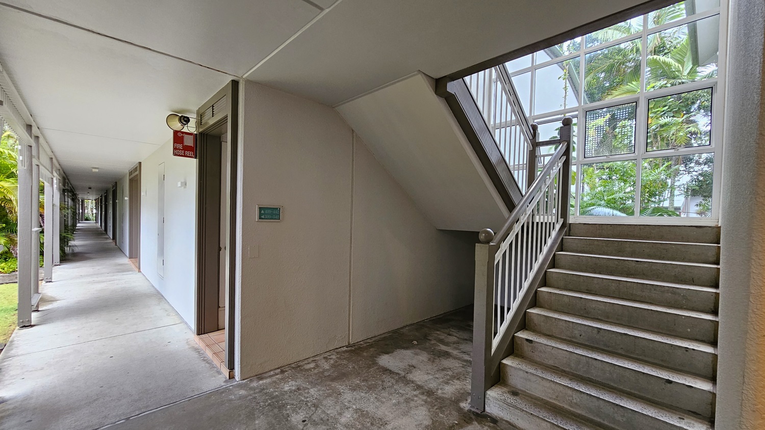On the left, a long corridor with beige walls and grey doors extends into the distance, with a fire hose reel mounted on the wall. To the right, a staircase with grey steps and white railings ascends next to a large window with a view of green palm trees. In the middle, the corridor and staircase meet at a corner. The floor is concrete, and the ceiling is flat and white with recessed lighting.