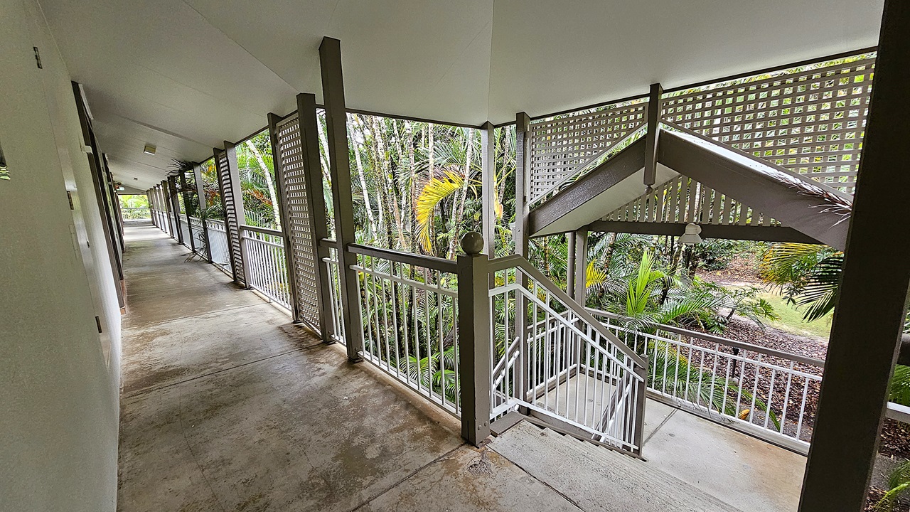 On the left, a long corridor with beige walls and a concrete floor extends into the distance, with doors and ceiling lights. To the right, a staircase with grey steps and white railings descends next to a lattice screen and green palm trees. In the middle, the corridor and staircase meet at a corner, with the staircase leading down to a garden area. The ceiling is flat and white with recessed lighting.