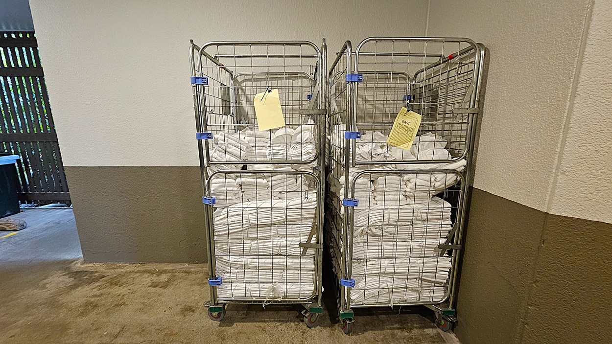On the left, a beige and grey wall with a wooden slatted fence in the background. To the right, two metal carts filled with neatly folded white towels are positioned against a beige and grey wall. In the middle, the carts have yellow tags attached to them.