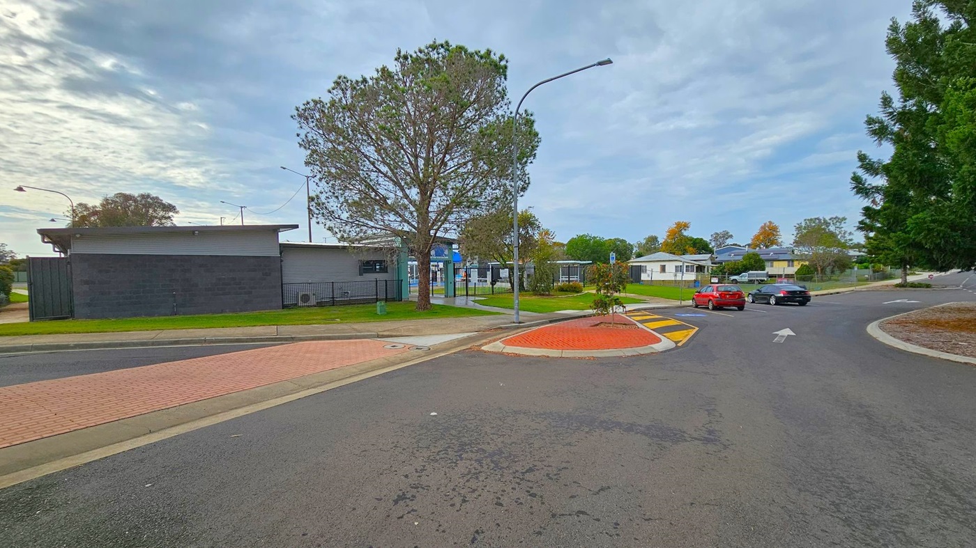 A street scene with a black building on the left, featuring a flat roof and surrounded by a grassy area. A large tree stands nearby, next to a metal fence. In the centre, a road curves to the right, with a red car and a black car driving away. On the right, there are more trees and a few residential buildings in the background. The road has a yellow and black speed bump and a red brick pedestrian crossing. Street lamps are visible along the road.