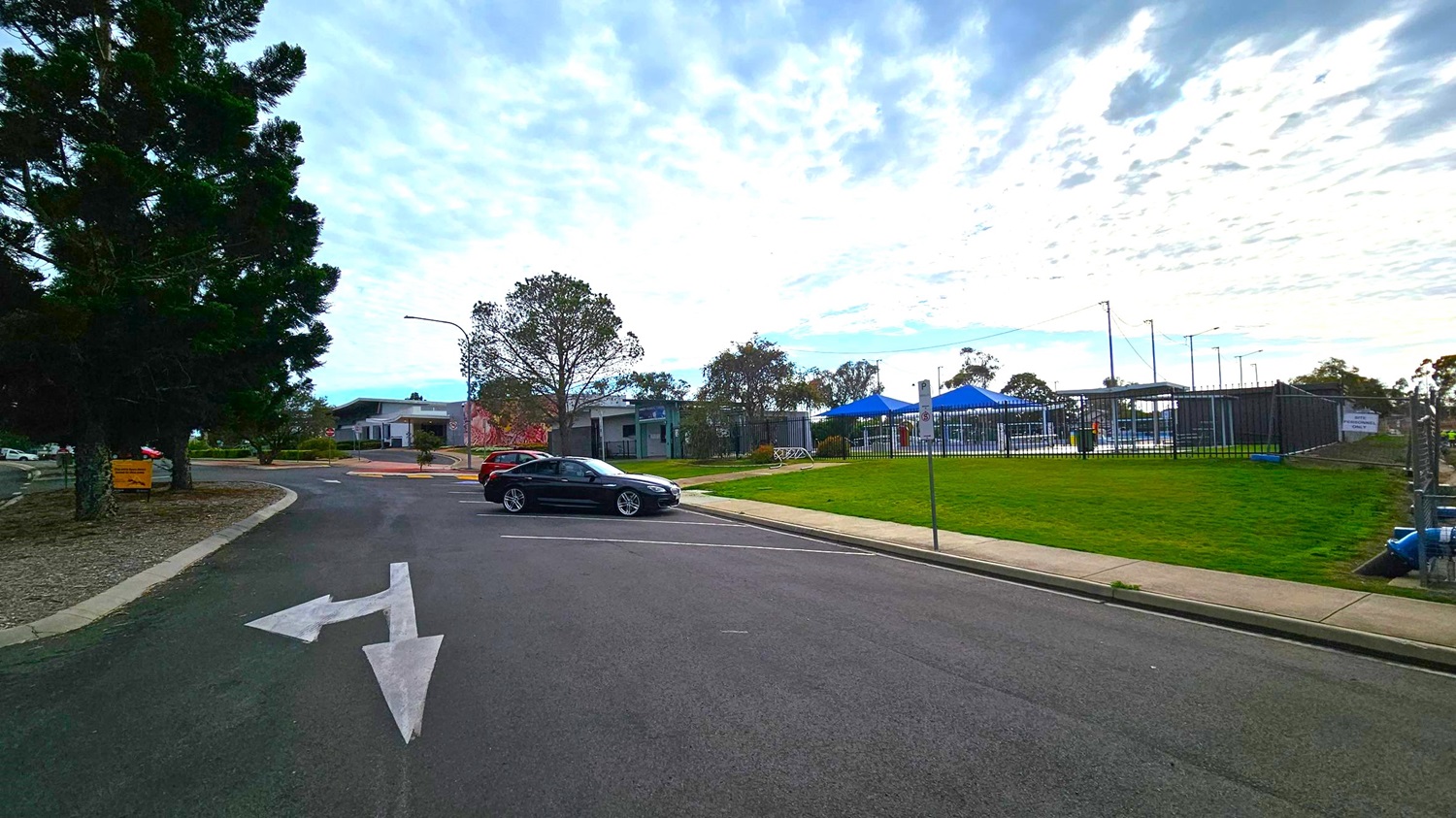 A road with a white directional arrow painted on it. On the left, there are tall trees and a small building in the distance. A black car is parked near the centre, with a grassy area and a fence to the right. Behind the fence, there are blue canopies and more trees. The sky is overcast with scattered clouds.