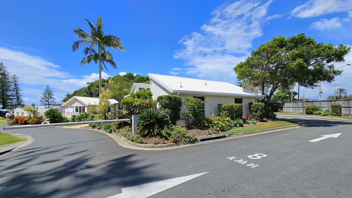 To the left, a concrete landmark displays the name "Ocean Beach Tourist Park" amid landscaped plants. The bitumen road splits, with a traffic barrier indicating no entry on the left. White markings guide travellers on the right path. Landscaped green grass fills the right side. Lamp posts and palm trees line the road. Low-set buildings with pitched roofs are positioned on the left on the left side of the road.