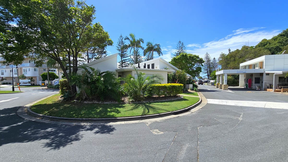 A roundabout surrounded by landscaped plants, trees, and grass. Behind it, a low-set building with a white concrete facade and pitched roof. On the right, another low-set building with a portico housing the reception office. To the left, the main road and another multi-storey building. Palm trees and pine trees are visible in the distance.