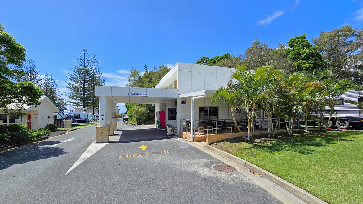 To the right is a low-set white building with a portico front. A ramp with handrails leads to the reception entrance, alongside tables and chairs. The road has markings denoting the entryway. On the left is another low-set building with a pitched roof and white concrete facade. Various trees and neatly trimmed grass are on the right. Further ahead, caravans are also visible.