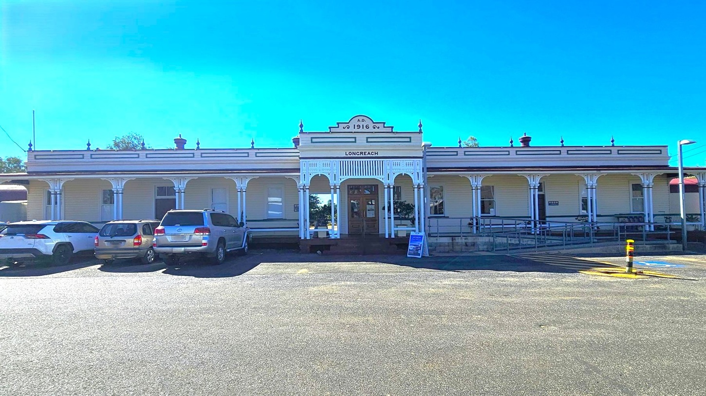 A historic building with a sign reading "LONGREACH" and "A.D. 1916" at the top. The structure features a series of arches and decorative columns along the front. Several cars are parked in front of the building on an asphalt surface. A small signboard is placed near the entrance. The building has a light-coloured facade with dark trim and a flat roof adorned with decorative elements. Trees are visible in the background.