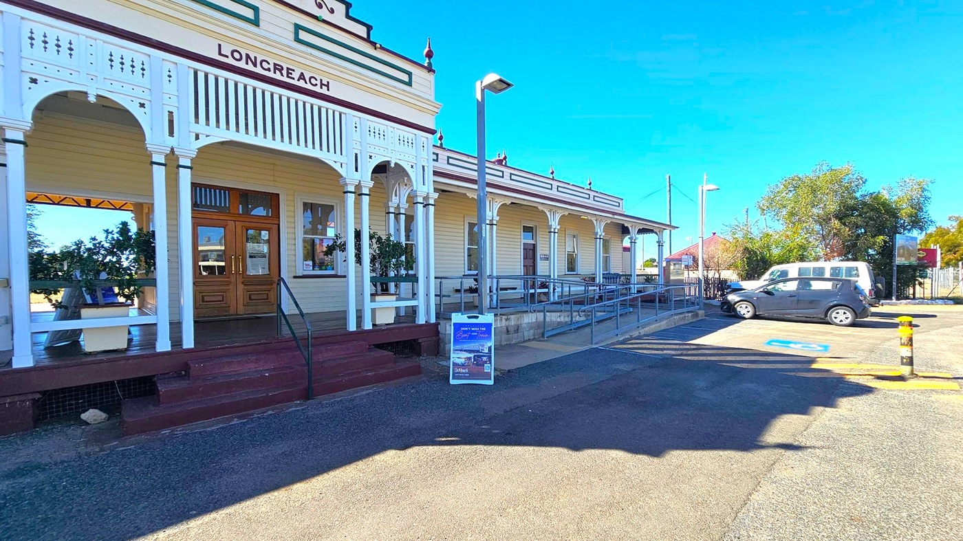 A historic building with a sign reading "LONGREACH" on the facade, featuring ornate white wooden arches and a wooden entrance door. A ramp and steps lead up to the entrance. In the foreground, a signboard is placed on the pavement. To the right, a parking area with two vehicles, one white and one black, is visible. Trees and a red-roofed building are in the background. The ground is paved, and a streetlamp is present near the ramp.