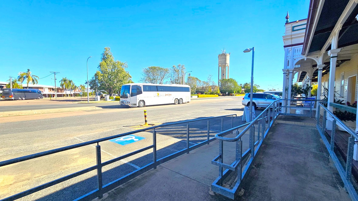 A street scene with a white tour bus parked on the left side of the road. In the background, there are palm trees, a large tree, and a tall water tower. On the right, a building with a covered walkway and decorative columns is visible. A silver car is parked nearby, and a ramp with metal railings leads up to the building. The pavement has a marked disabled parking space. The sky is clear and blue.