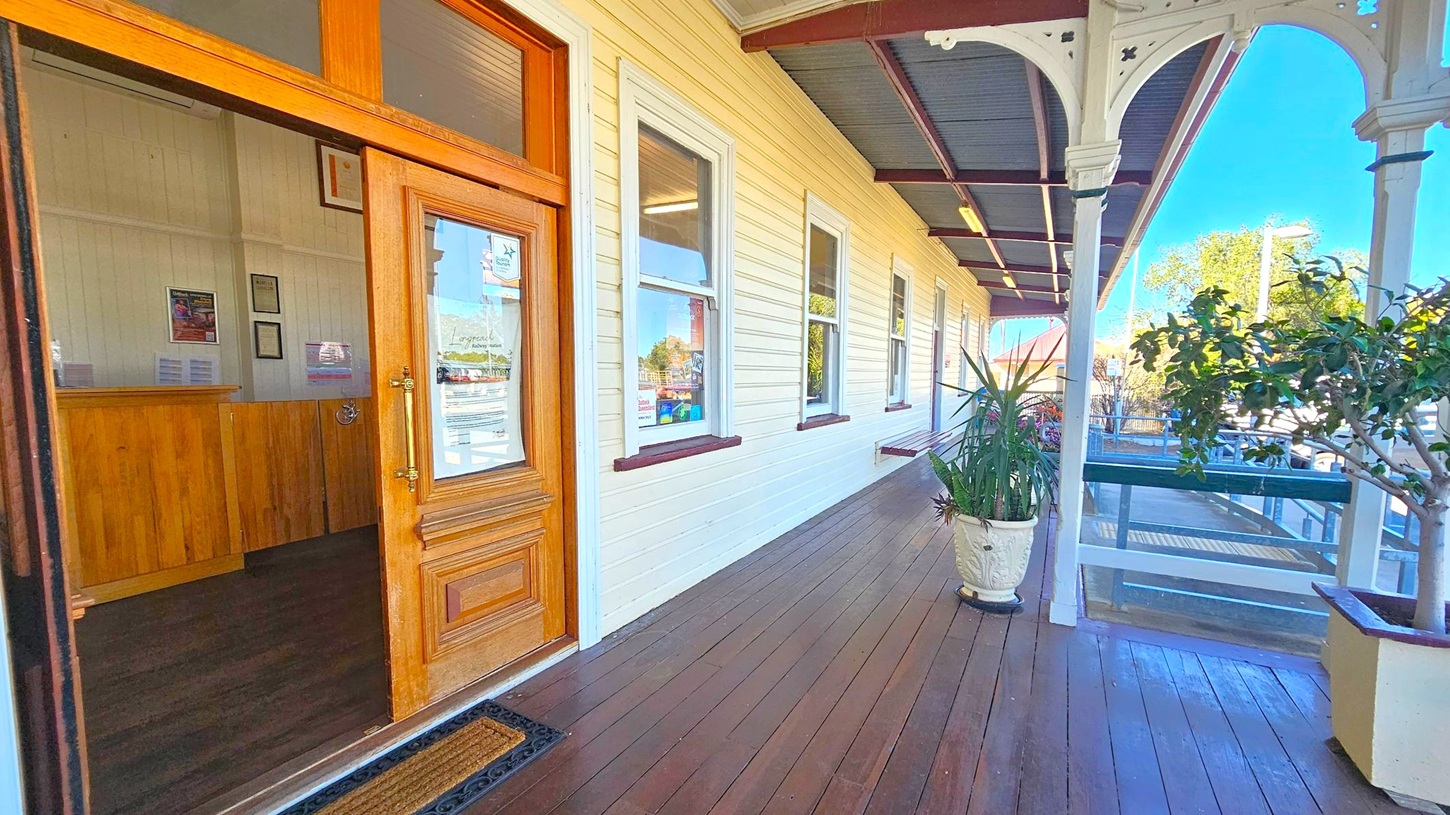 A wooden porch with a polished floor extends along the side of a building. On the left, a wooden door is open, revealing an interior with framed items on the wall. The building's exterior is light-coloured with several windows. On the right, potted plants are placed along the porch, and decorative wooden posts support the overhanging roof. In the background, a ramp leads down, and trees and a building with a red roof are visible.