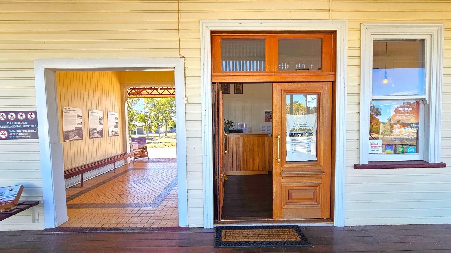 A wooden building with cream-coloured walls. On the left, an open entrance leads to a room with tiled flooring and informational posters on the walls. A bench runs along the wall. In the centre, a wooden door with glass panels has a notice attached. To the right, a window displays various posters and brochures. Outside, trees and a clear view of the sky are visible through the open entrance. A doormat lies in front of the door.