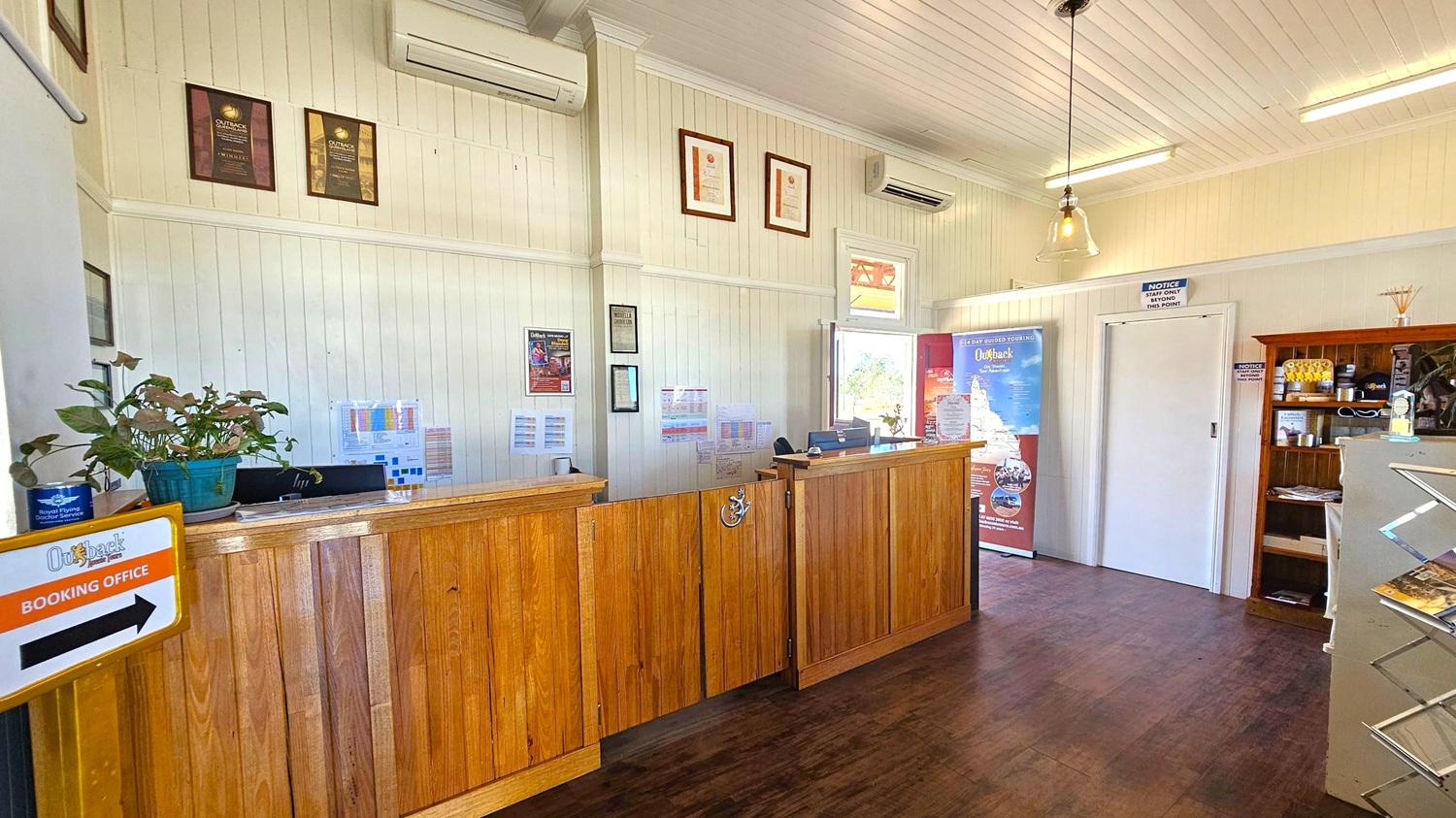 A reception area with a wooden counter on the left, adorned with a potted plant and a sign indicating a booking office. The walls are light-coloured with several framed certificates and awards. An air conditioning unit is mounted above. On the right, there's a door, a promotional stand, and a wooden shelf with various items. The floor is dark wood, and the ceiling is white with pendant and fluorescent lights. A window in the background offers a view outside.