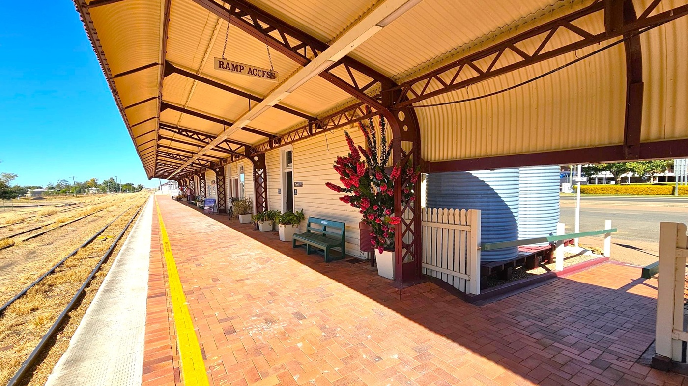 A railway platform with a covered walkway on the right, featuring a corrugated iron roof supported by metal beams. The platform is paved with red bricks and has a yellow safety line near the edge. A sign reads "RAMP ACCESS." There are benches and potted plants along the wall of the building. On the far right, a large water tank is visible behind a white picket fence. The railway tracks run parallel to the platform on the left, with dry grass between them.