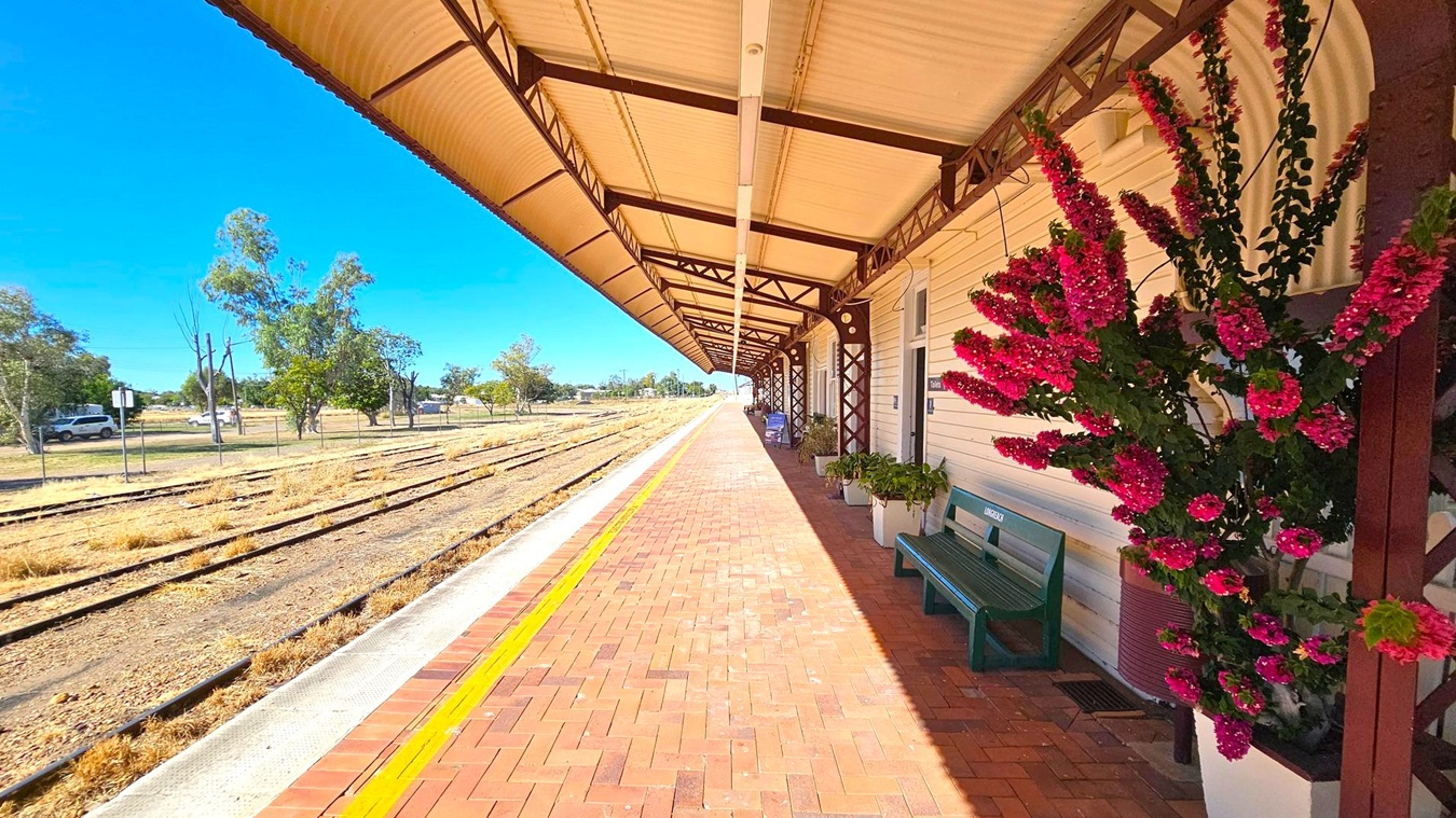 A railway station platform with a long, covered walkway. On the right, there is a building with a wooden exterior, adorned with vibrant pink flowers in planters. A green bench is positioned against the wall. The platform is paved with red bricks and has a yellow safety line near the edge. On the left, multiple railway tracks run parallel, bordered by dry grass. In the background, there are scattered trees and a few parked vehicles. The ceiling is supported by metal beams.